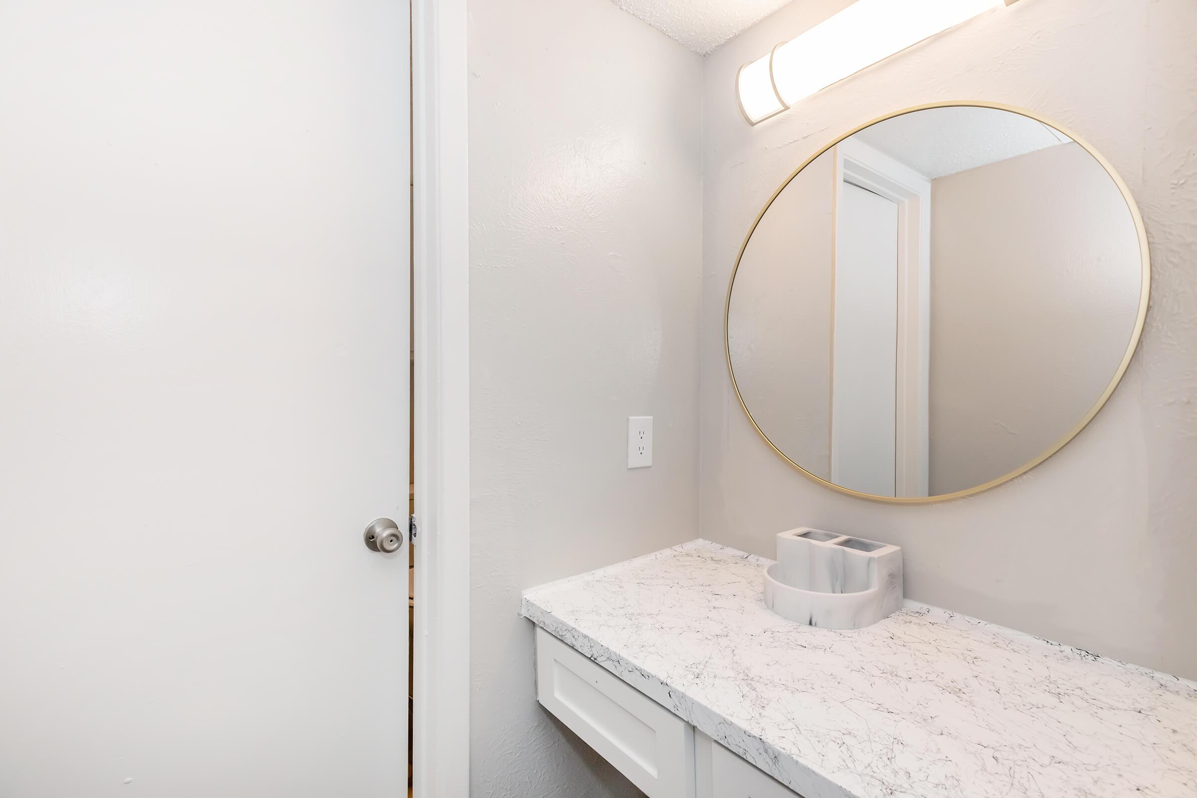 A modern bathroom setting featuring a round mirror with a gold frame above a white marble countertop. The countertop has a white container for toiletries. A white door is partially visible, and the walls are painted in a light gray color. A light fixture is mounted above the mirror, providing illumination.