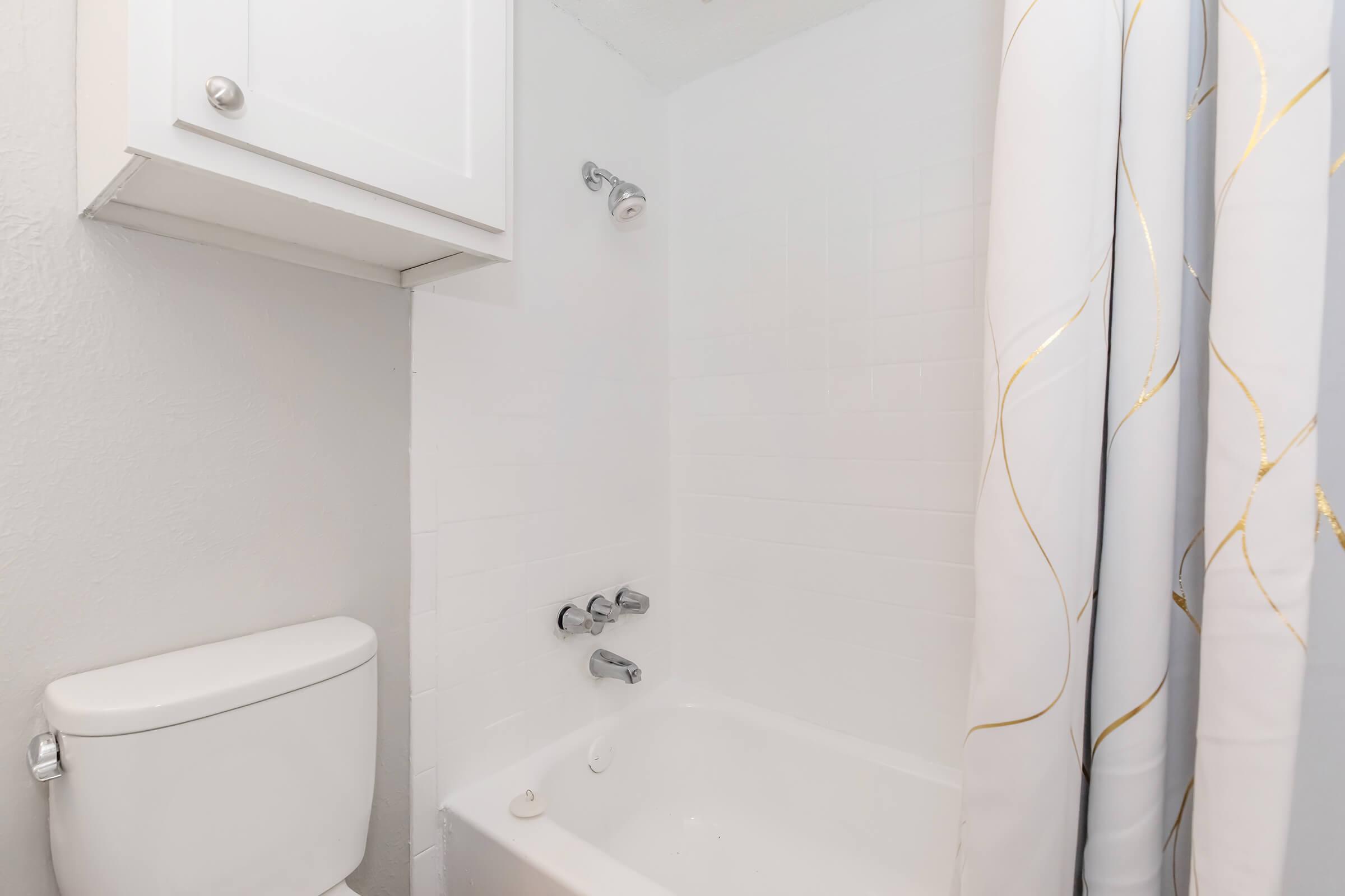 A clean bathroom featuring a white bathtub with a silver faucet and showerhead, a white toilet, and a white cabinet above the tub. The shower has a decorative curtain with a gold and white design. The walls are plain and light-colored, contributing to a bright and fresh ambiance.