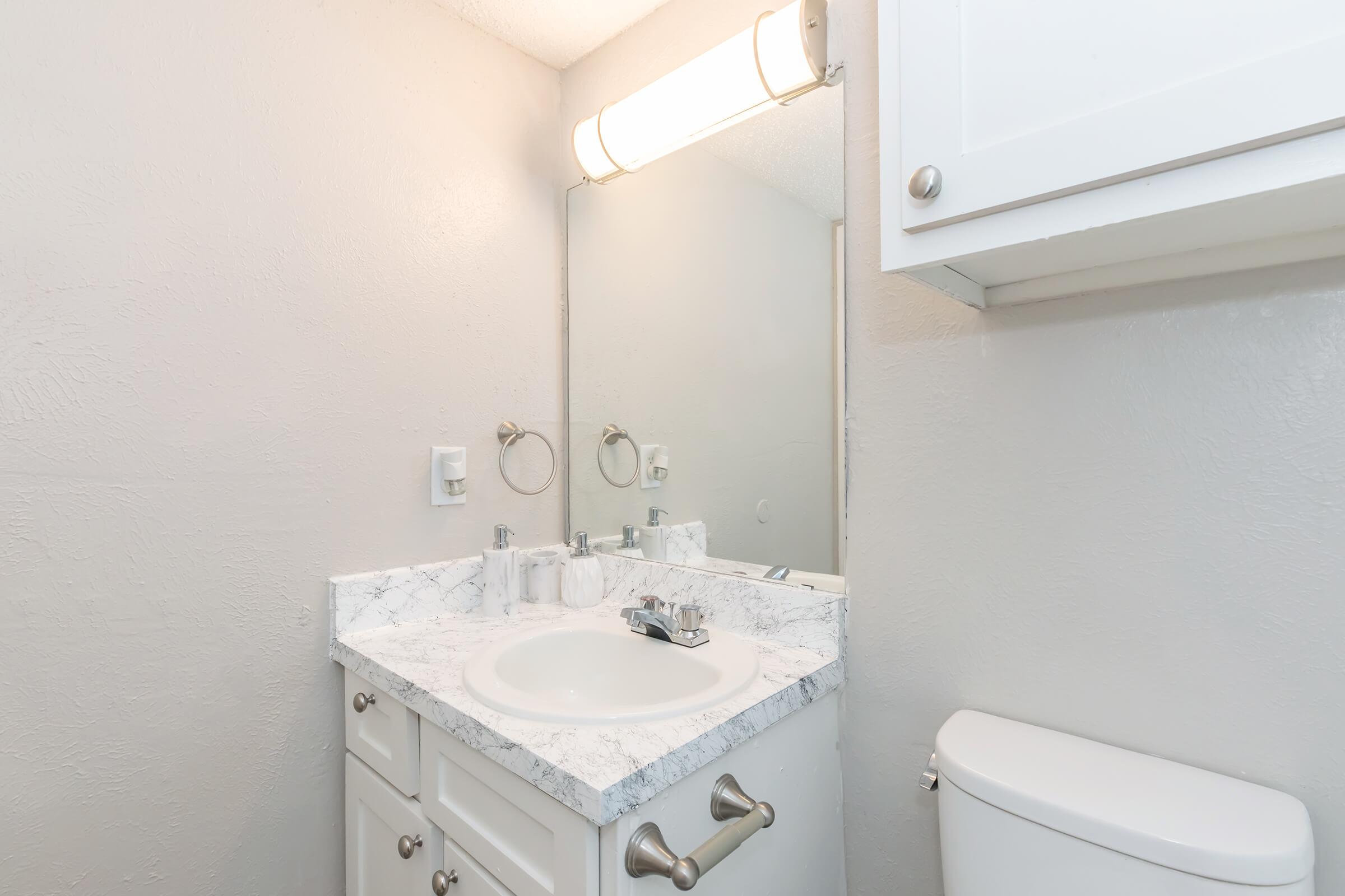 A modern bathroom featuring a marble countertop sink with a round mirror above it. There are silver faucet fixtures, two wall-mounted circular mirrors, and a white toilet. The walls are painted in a light shade, and there is a white cabinet above the sink for storage. Soft lighting is provided by a wall-mounted light fixture.