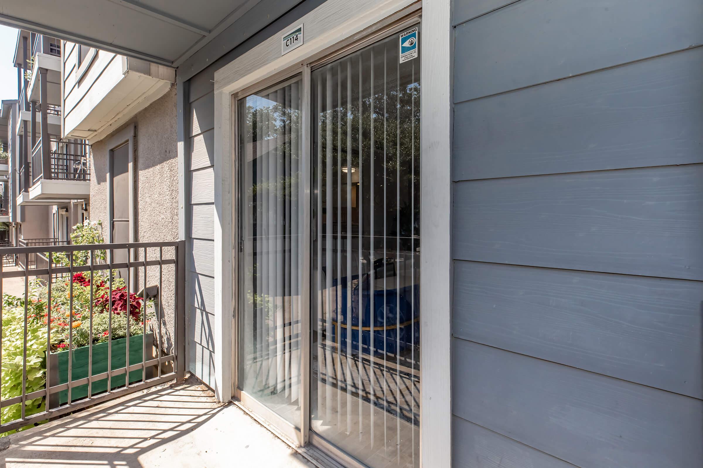 A view of a modern apartment entrance with sliding glass doors, featuring vertical blinds. The exterior wall is painted in light gray, and there is a small railing in front of a flowerbed with colorful plants. Nearby, part of another building is visible, suggesting an outdoor living space.