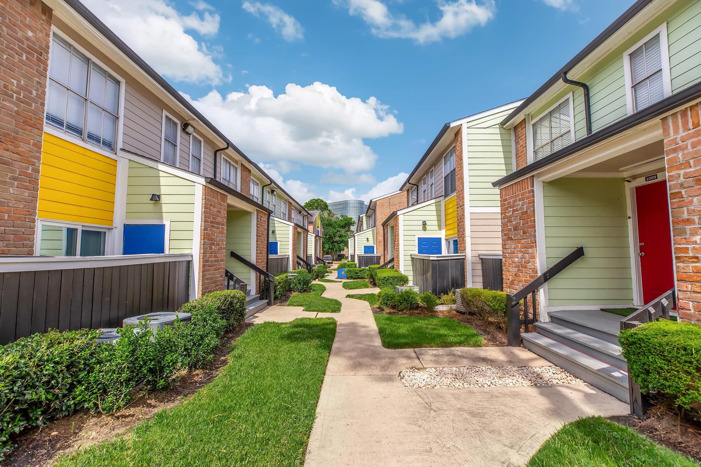 A landscaped pathway between two rows of residential buildings with colorful façades. The apartments feature a mix of brick and light-colored siding, with bright yellow and blue accents. Lush green grass and neatly trimmed bushes flank the walkway, under a partly cloudy blue sky.
