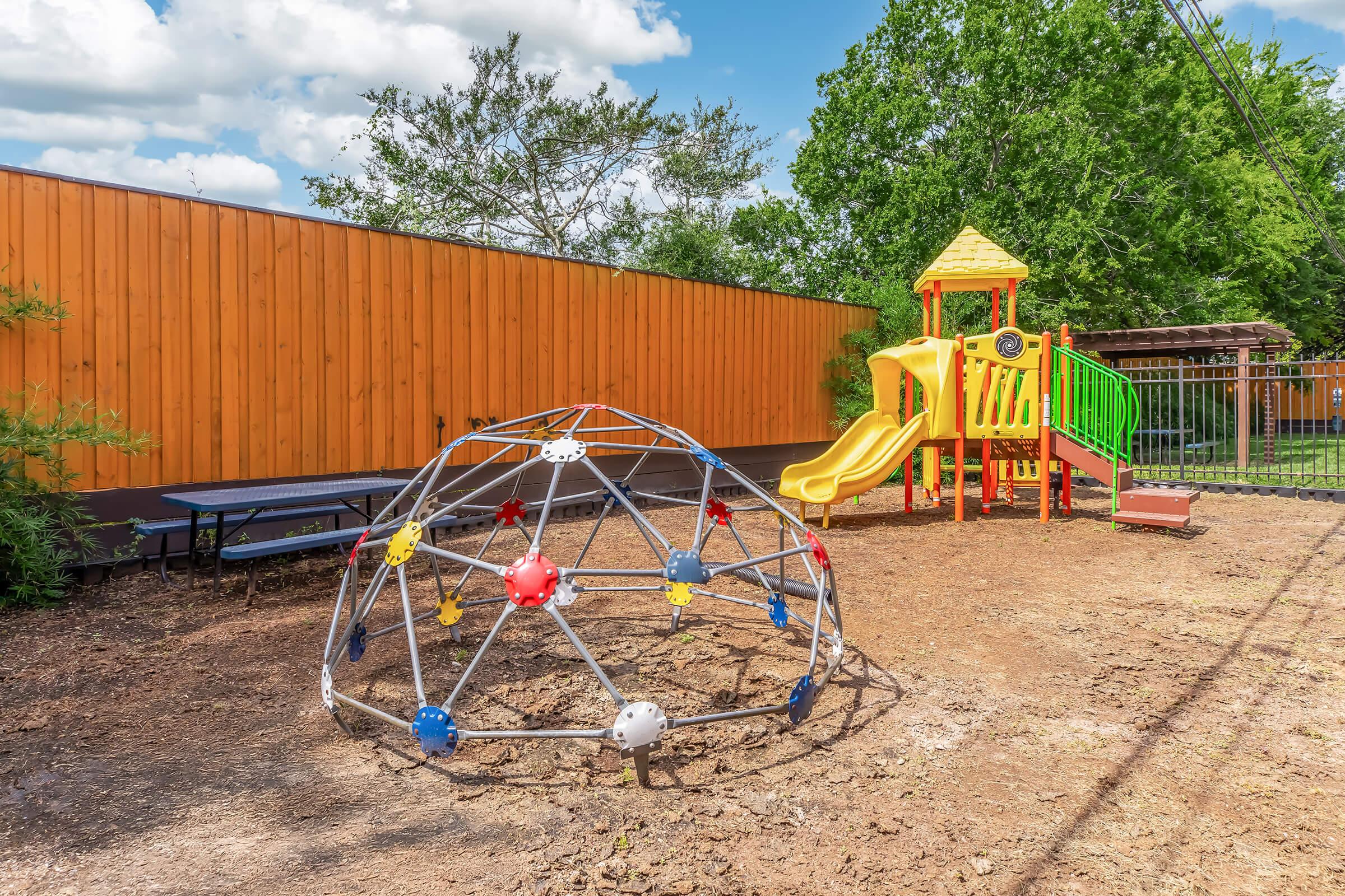 A colorful playground featuring a slide and climbing structure, alongside a geodesic climbing dome. The area is surrounded by greenery and has a wooden fence in the background, with bright blue skies and fluffy clouds overhead. The ground is dirt, creating a natural play environment.