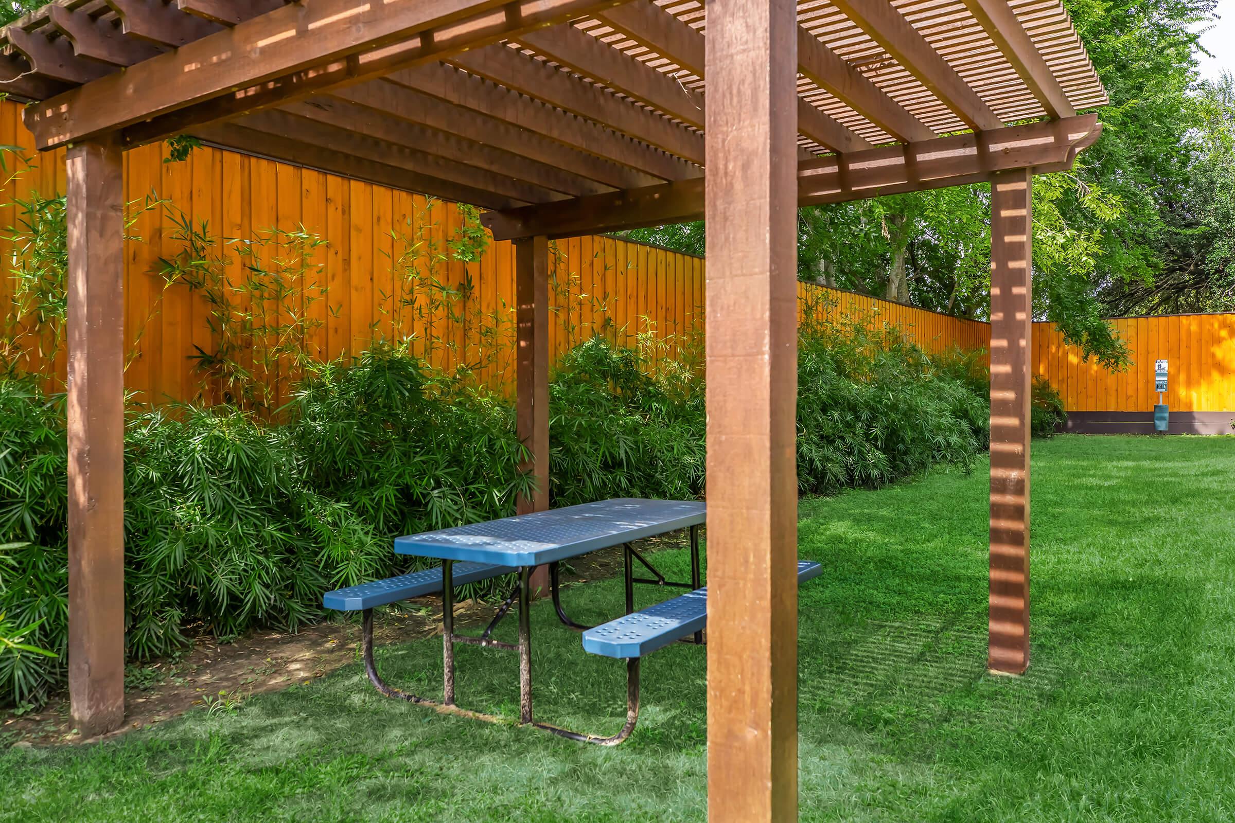 A wooden pergola with a blue picnic table underneath, surrounded by lush green grass and plants. In the background, there is a bright orange wooden fence, adding a vibrant contrast to the greenery. The setting offers a cozy outdoor space for relaxing or dining.