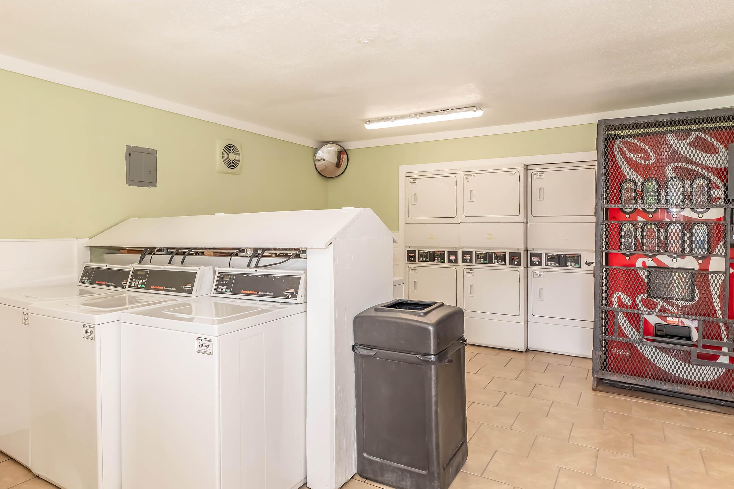 A clean laundry room featuring multiple white washing machines and dryers, a black trash can, and vending machines behind a metal cage. The walls are painted a light green, and there are fluorescent lights providing bright illumination. The space appears well-maintained and functional for laundry tasks.