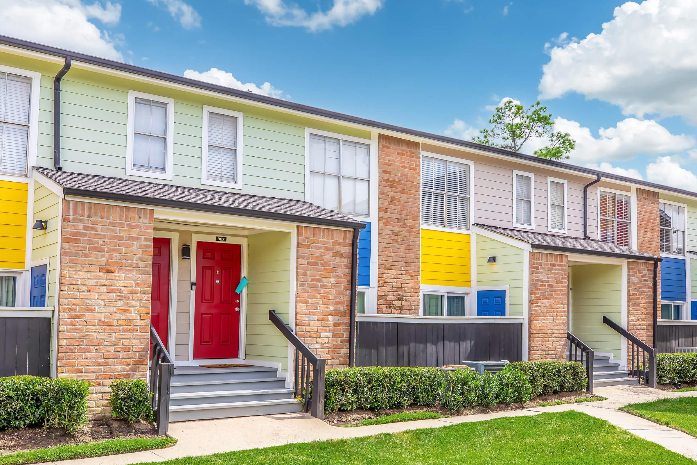 Colorful townhouse exteriors with a mix of brick and painted wooden facades. The first unit features a bright red door, while other units have blue and yellow accents. Neatly trimmed shrubs line the pathways, and the sky is partly cloudy, creating a cheerful suburban atmosphere.