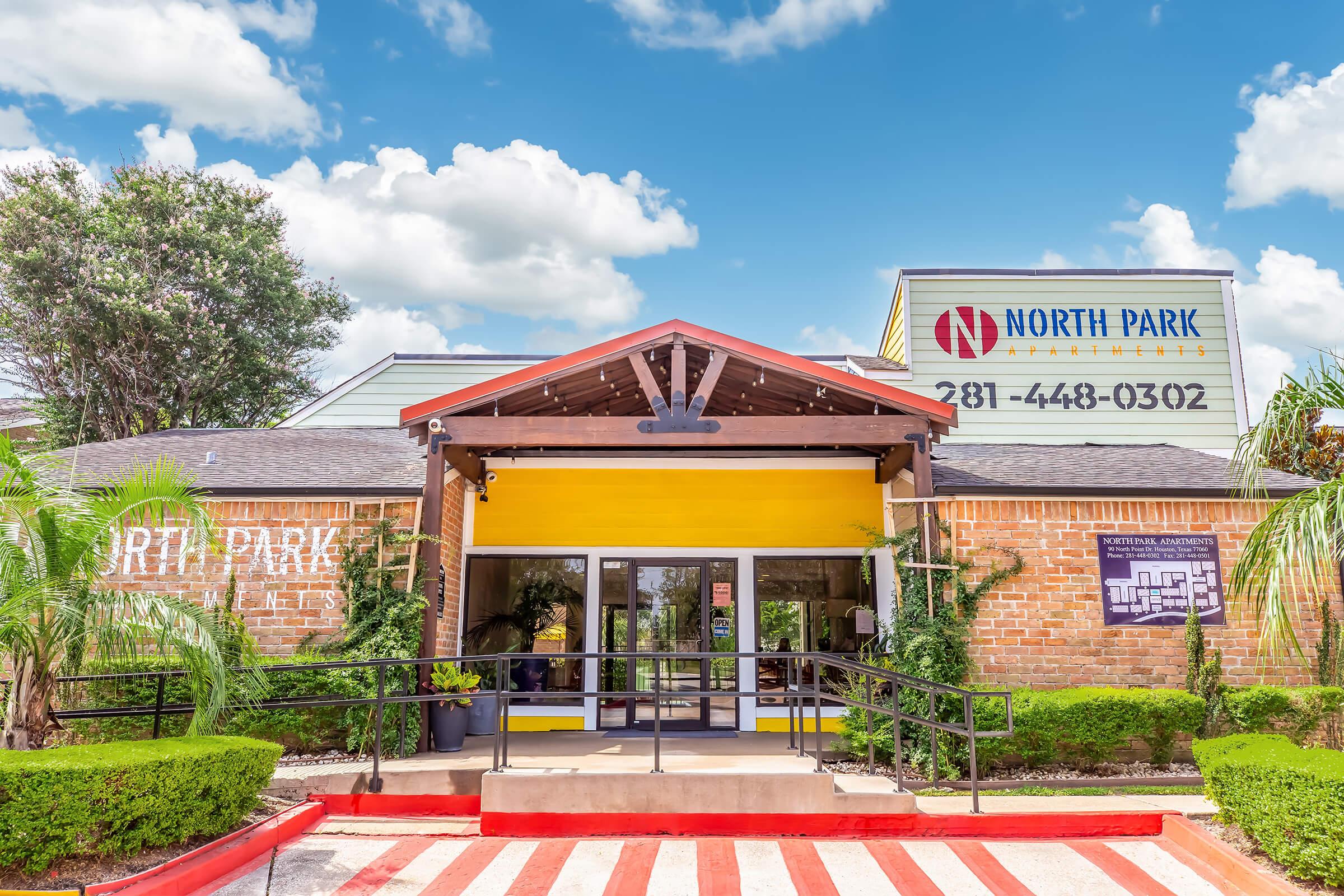 Exterior view of North Park Apartments with a brick façade and a yellow entrance awning. The entrance is flanked by greenery, and a red-striped pathway leads to the door. Above the entrance, there's a sign displaying the name and contact number. The sky is bright blue with fluffy white clouds.