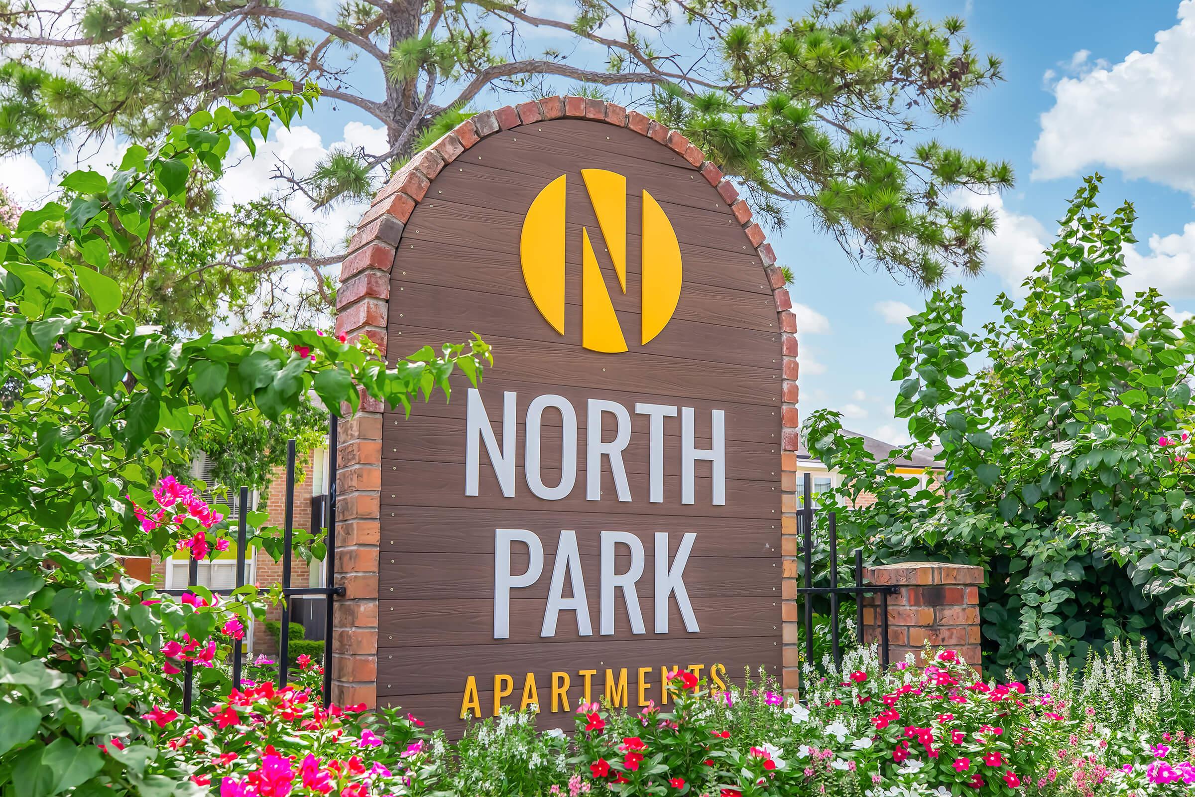 Sign for North Park Apartments surrounded by vibrant flowers and greenery. The sign features a large yellow "N" logo above the name "NORTH PARK" and the word "APARTMENTS" in bold letters. The background includes a clear blue sky with scattered clouds, creating a welcoming atmosphere.