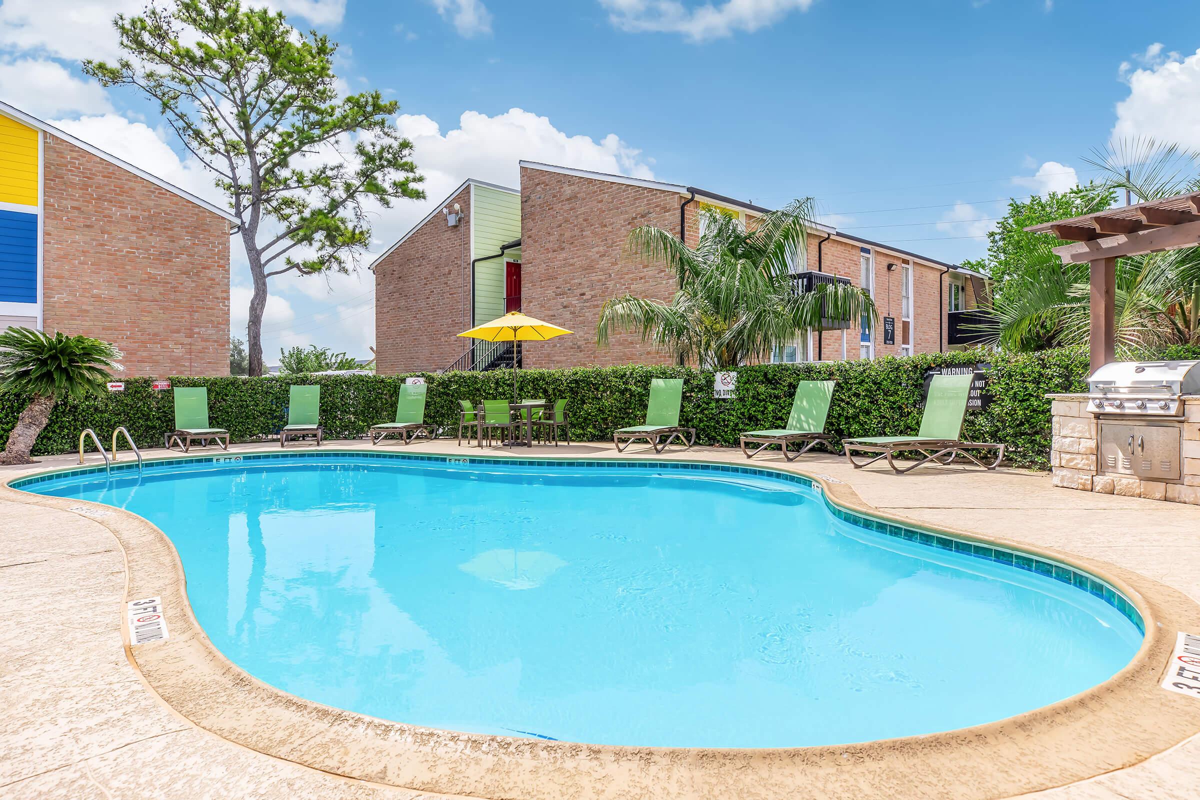A clear blue swimming pool surrounded by green lounge chairs and a small table with an umbrella. In the background, there are red brick apartment buildings and lush greenery, creating a relaxing outdoor atmosphere.