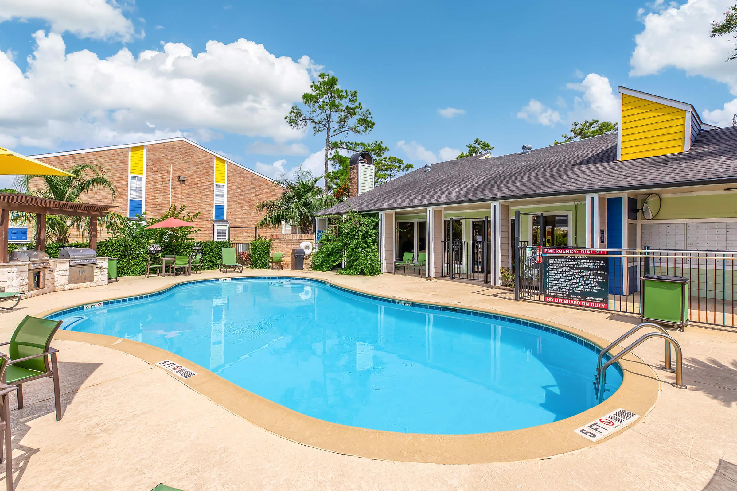 A well-maintained outdoor swimming pool surrounded by lounge chairs and tables, with a colorful apartment building in the background. The sky is bright with a few clouds, and there are palm trees and greenery around the pool area, creating a welcoming and relaxing atmosphere.