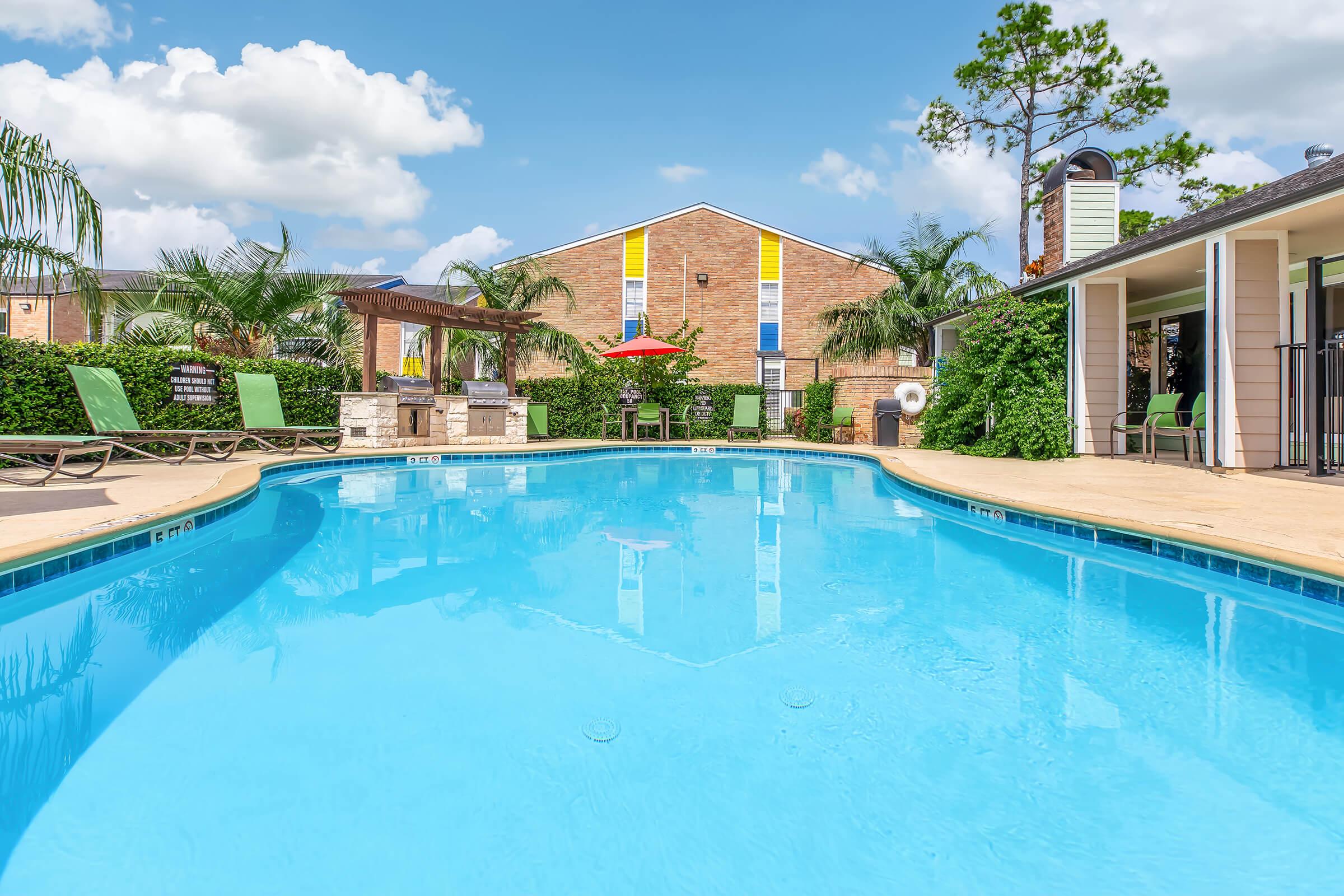A bright outdoor swimming pool surrounded by lounge chairs and tropical plants. In the background, a brick building with colorful accents is partially visible, along with a shaded seating area featuring a red umbrella. The sky is clear with fluffy white clouds.