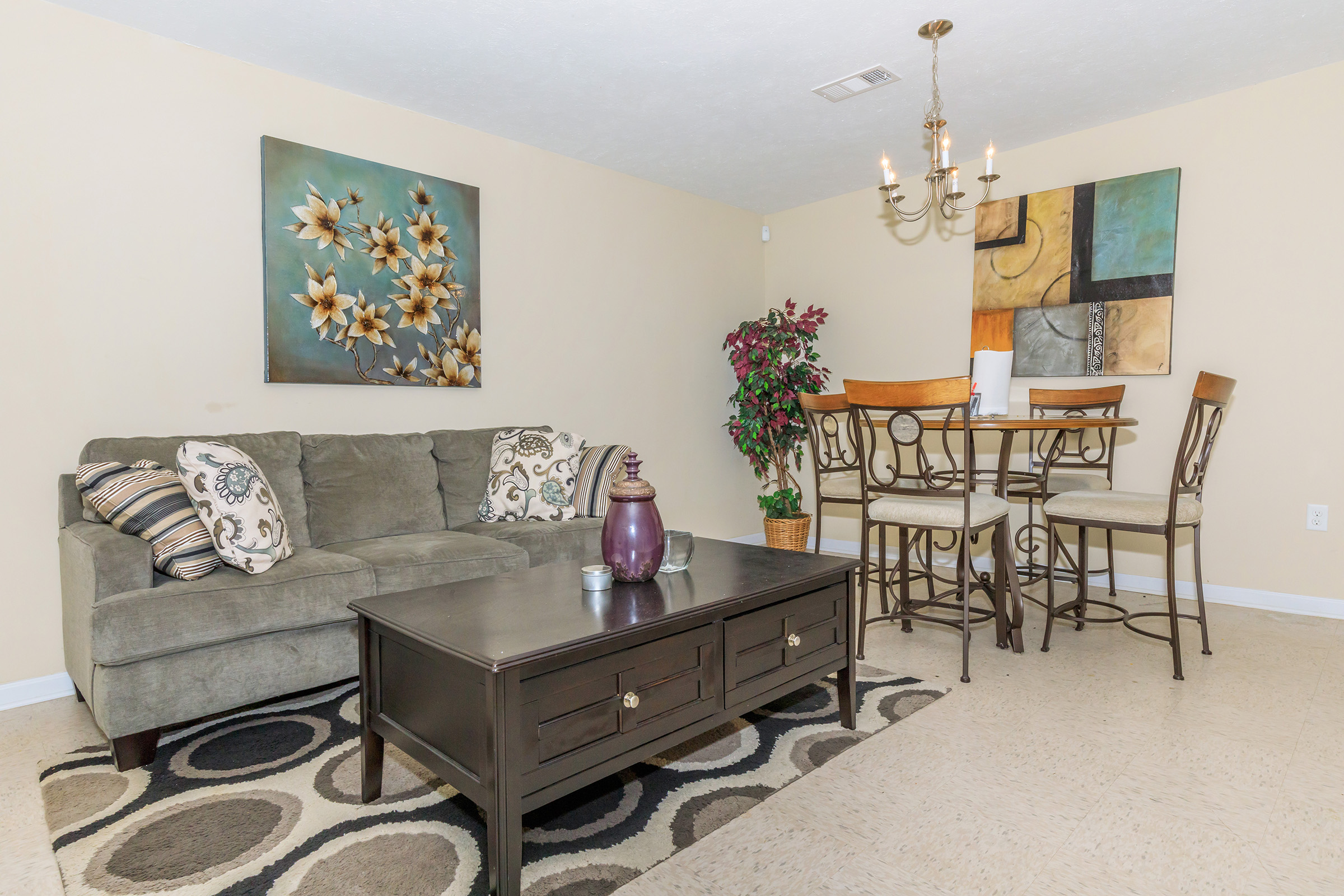 A cozy living space featuring a gray sofa with decorative pillows, a dark coffee table, and a dining area with a tall table and chairs. The walls are adorned with art, including a floral piece. A potted plant adds a touch of greenery, and the floor has a patterned rug for added warmth.
