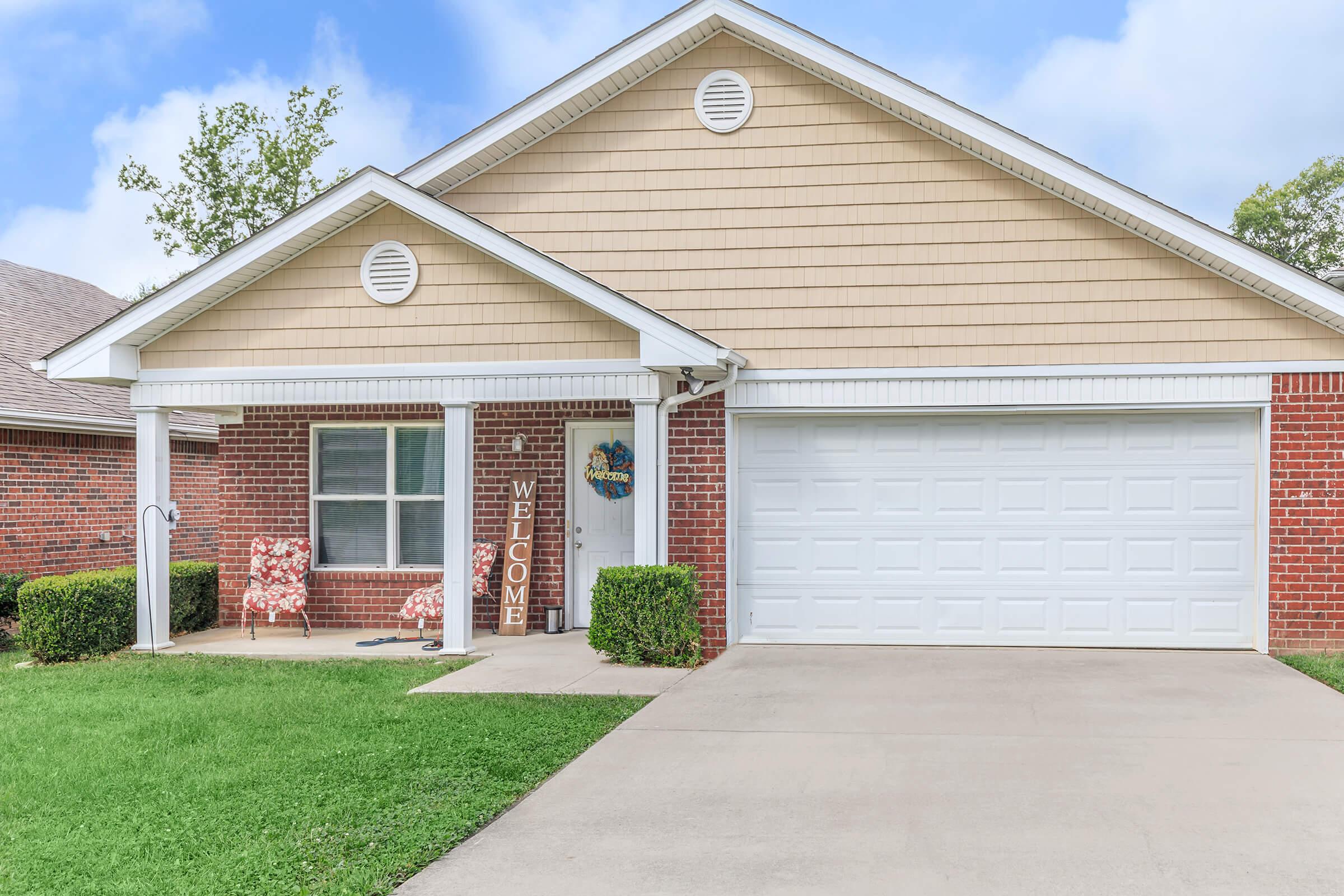 A well-maintained single-story house with beige siding and brick accents. The front features a welcoming porch with two red-patterned chairs, a "WELCOME" sign, and a neatly trimmed green lawn. A two-car garage is visible, along with a clear blue sky in the background.