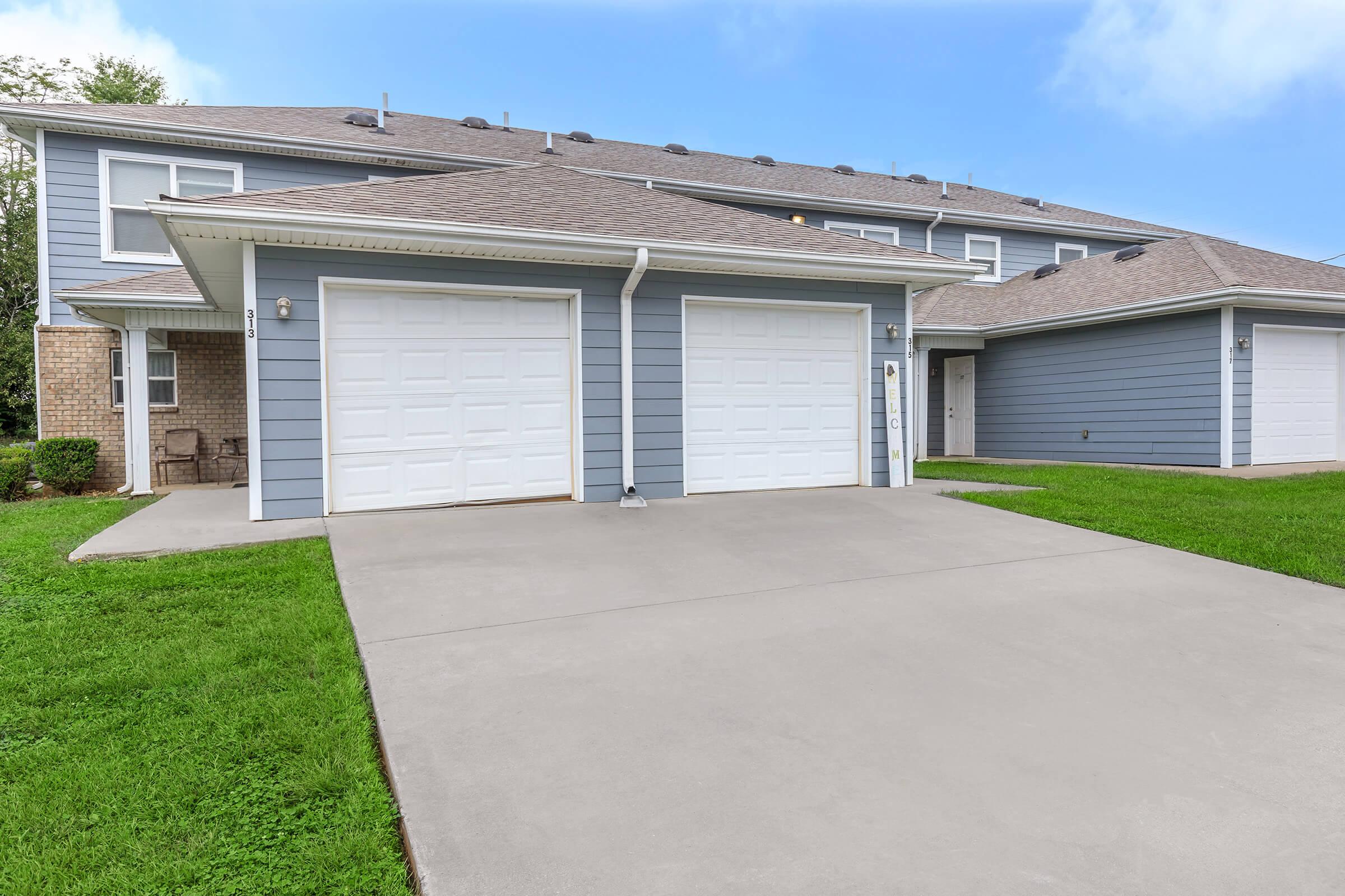 A modern two-car garage with white garage doors, set in front of a blue and beige multi-family building. The driveway is made of concrete and is surrounded by green grass, with a clear blue sky in the background.