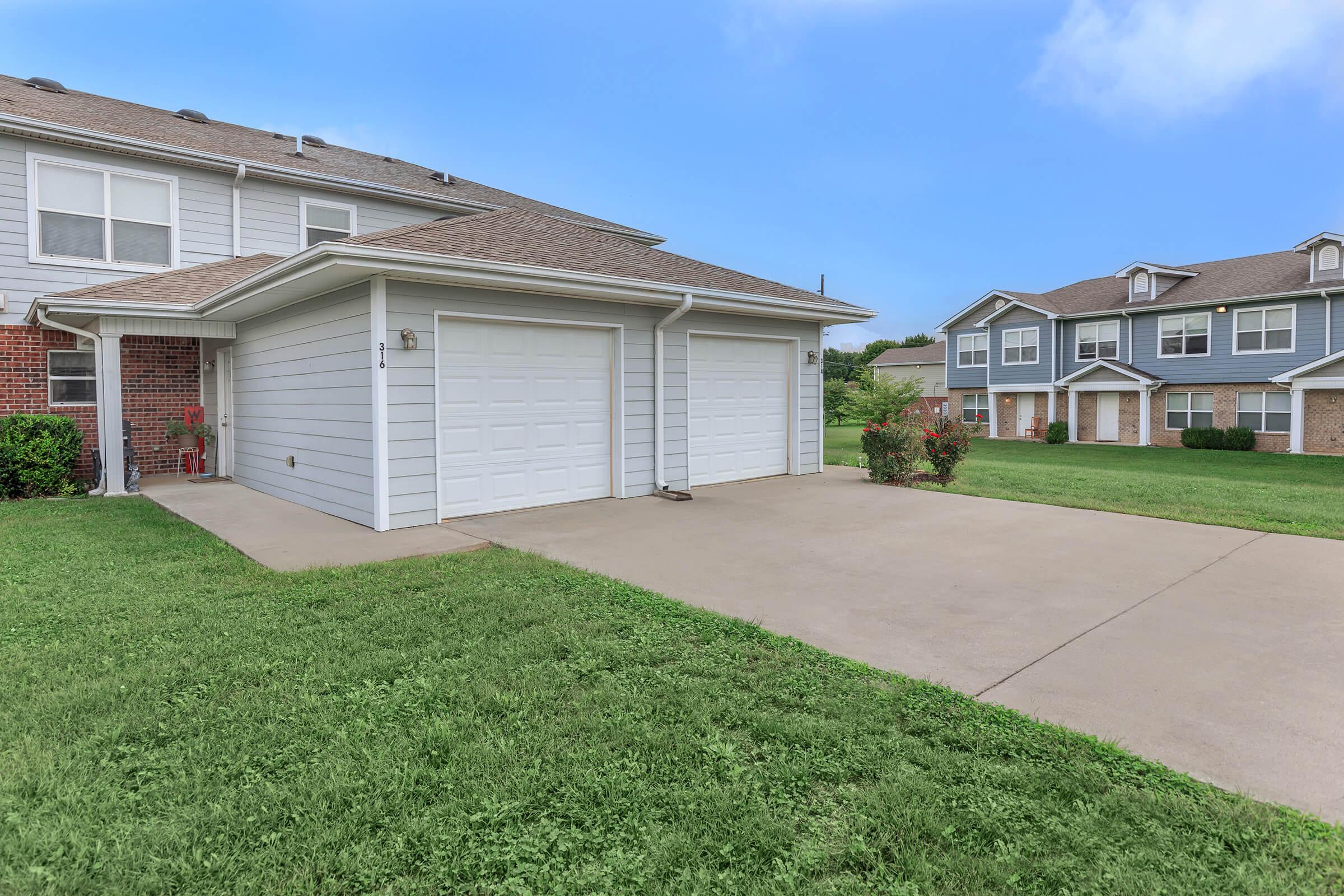 A well-maintained residential property featuring a two-car garage with a concrete driveway, surrounded by green grass. The building is two stories high with a light gray exterior and includes neighboring homes visible in the background under a clear blue sky.
