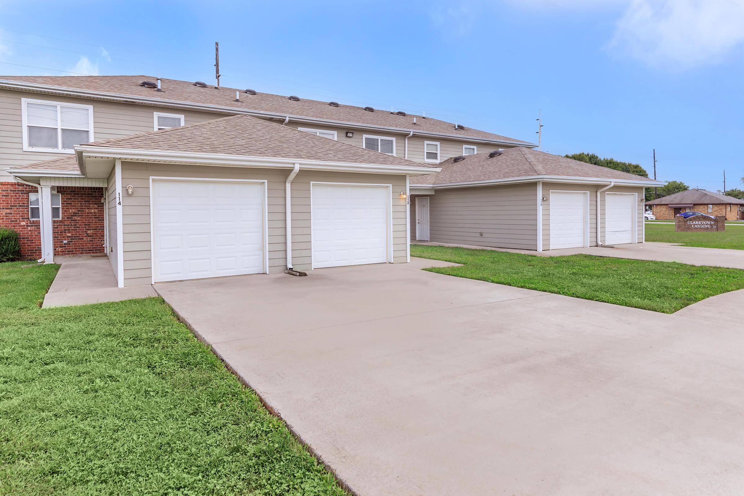 Two garages with white doors situated next to a light-colored residential building. The area features a paved driveway and green grass, with a clear blue sky in the background. The scene conveys a clean and well-maintained suburban environment.