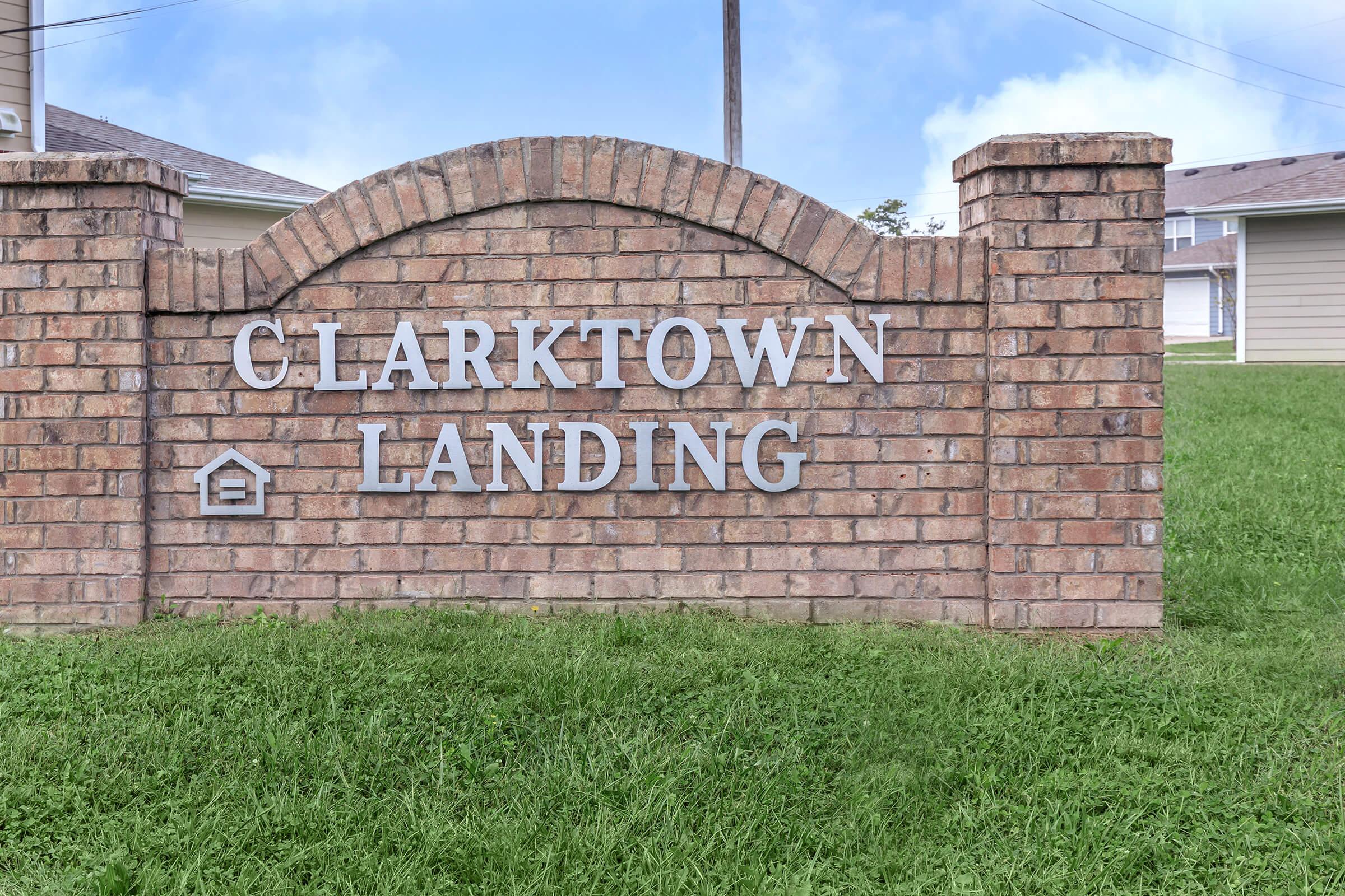 Brick sign displaying the words "CLARKTOWN LANDING" with a small house icon, set against a green grassy background. The sign is surrounded by a curved brick wall. In the background, there are residential buildings and a clear blue sky.