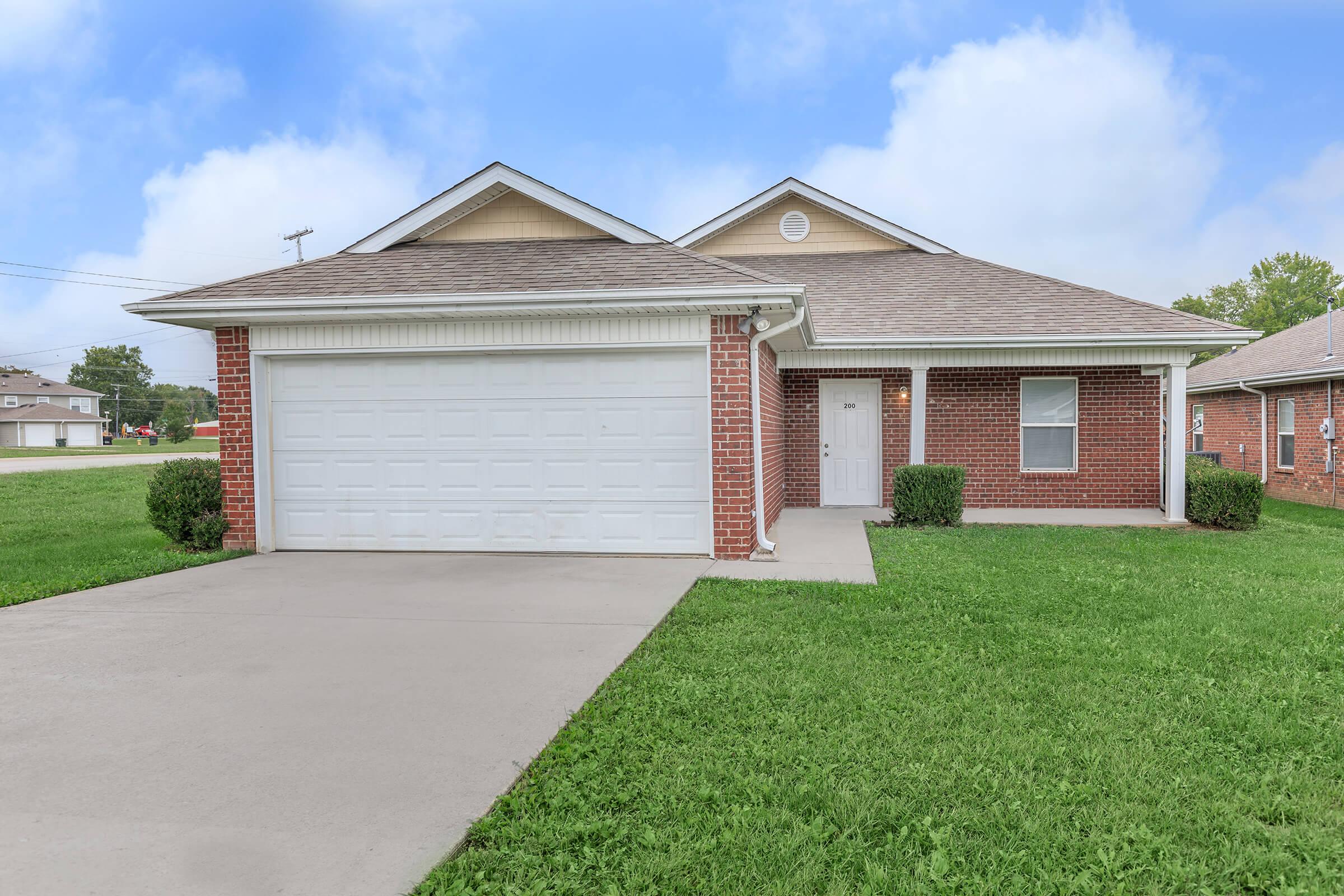 A single-story brick house with a front porch, a two-car garage, and a well-maintained lawn. The house features a neutral-colored exterior and is set against a clear blue sky with a few clouds in the background.