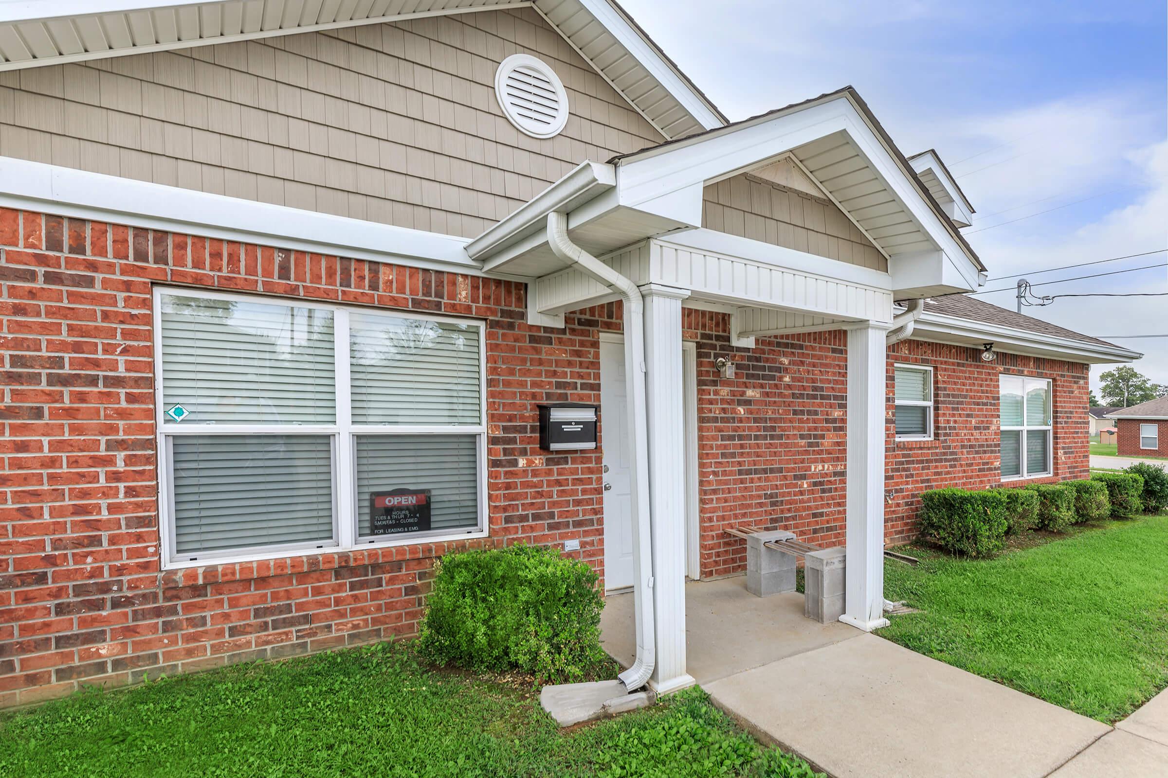 A brick building with a covered entrance, featuring a front porch and two large windows. The window displays an "OPEN" sign, and there are neatly trimmed shrubs along the walkway. The sky is partly cloudy, with green grass surrounding the area.