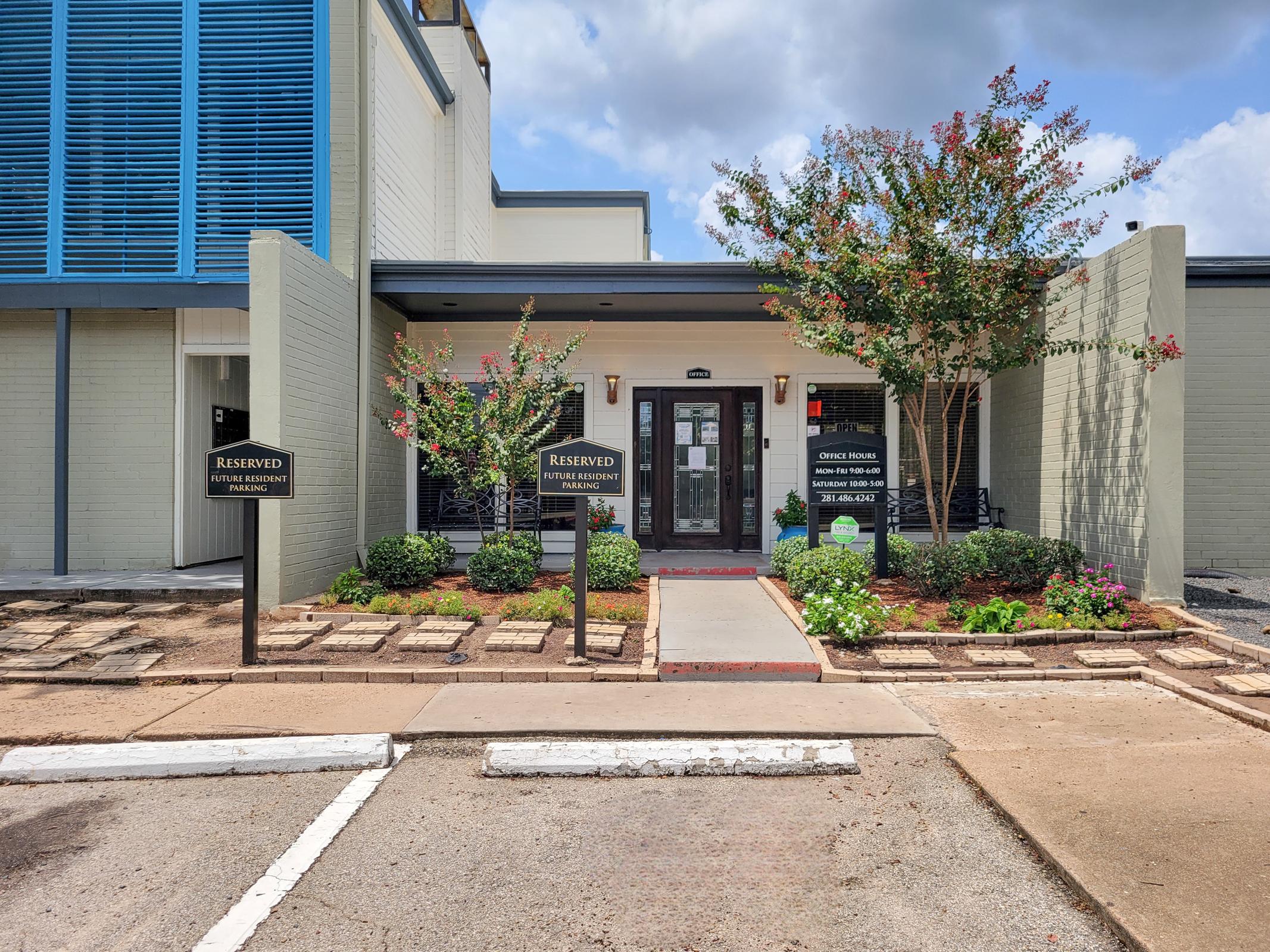 A modern building entrance with a landscaped pathway, featuring decorative plants and reserved parking signs. The entryway is framed by gray walls and blue accents, with a welcoming door and large windows. Above, the sky is partially cloudy, enhancing the inviting atmosphere of the location.