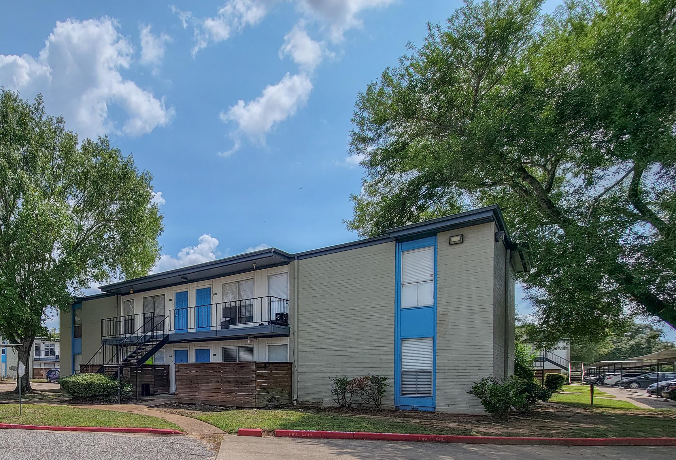 A two-story apartment building with light gray walls and blue doors. The building is surrounded by greenery, including large trees and well-maintained grass. The sky is partly cloudy with visible blue patches, and a walkway leads up to the entrance of the building.