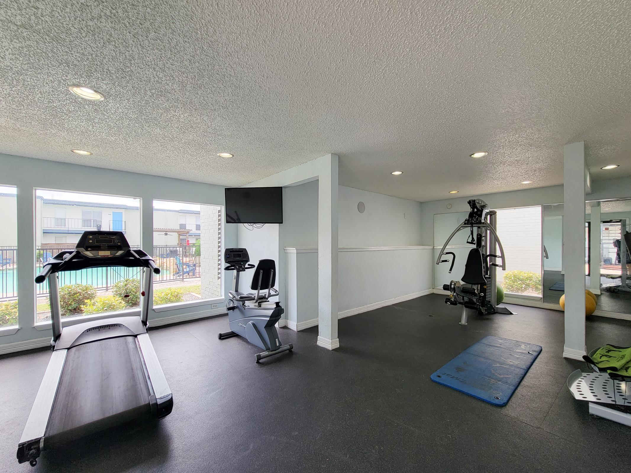 A bright and spacious gym with large windows showing a pool outside. It features a treadmill, a stationary bike, and a cable machine. The flooring is dark, and the walls are light-colored, contributing to an inviting atmosphere for exercise.