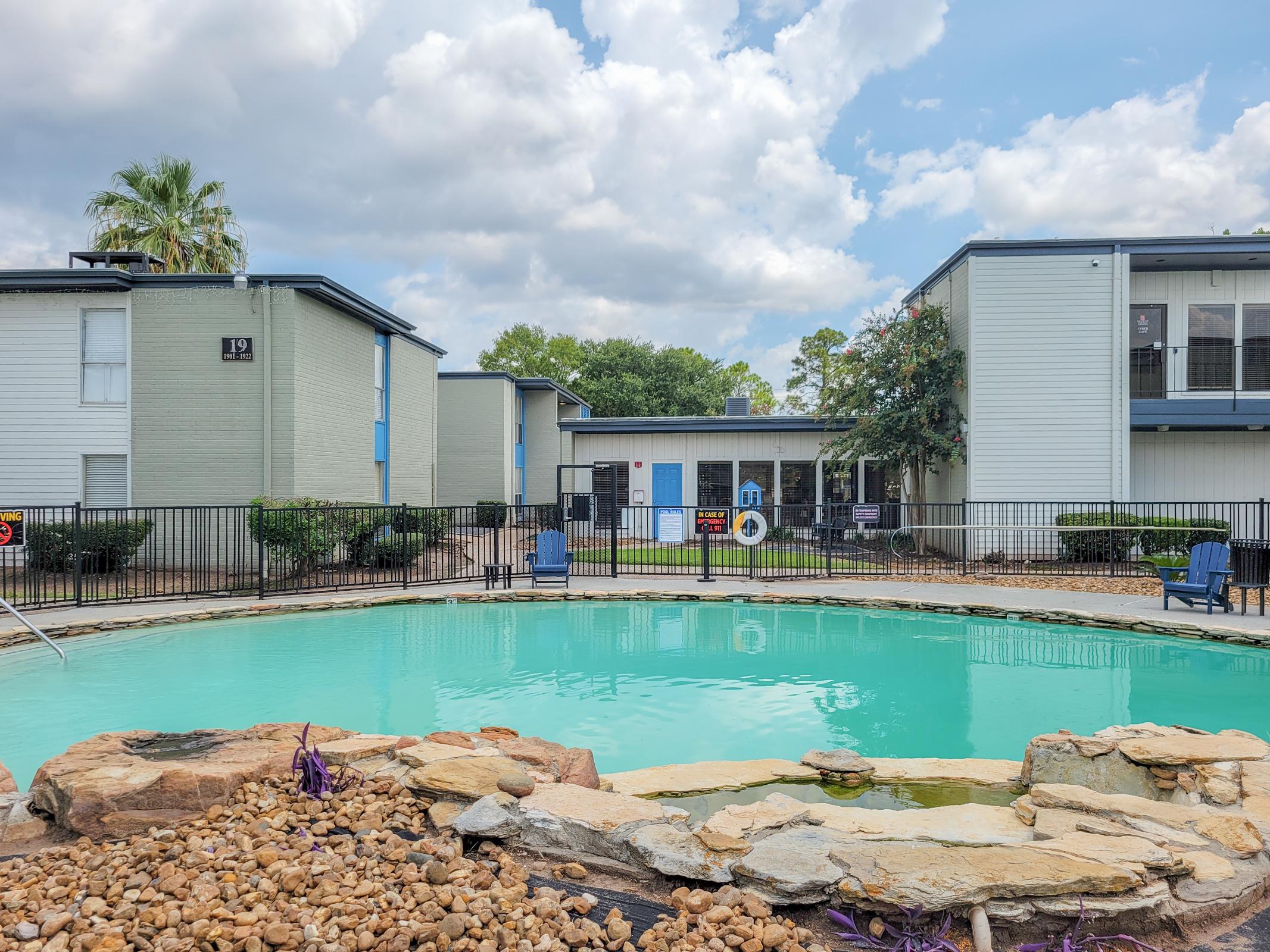 A tranquil swimming pool surrounded by a stone border, with lush landscaping and palm trees in the background. Two residential buildings with blue accents are visible on either side of the pool. The sky is partly cloudy, creating a serene atmosphere.