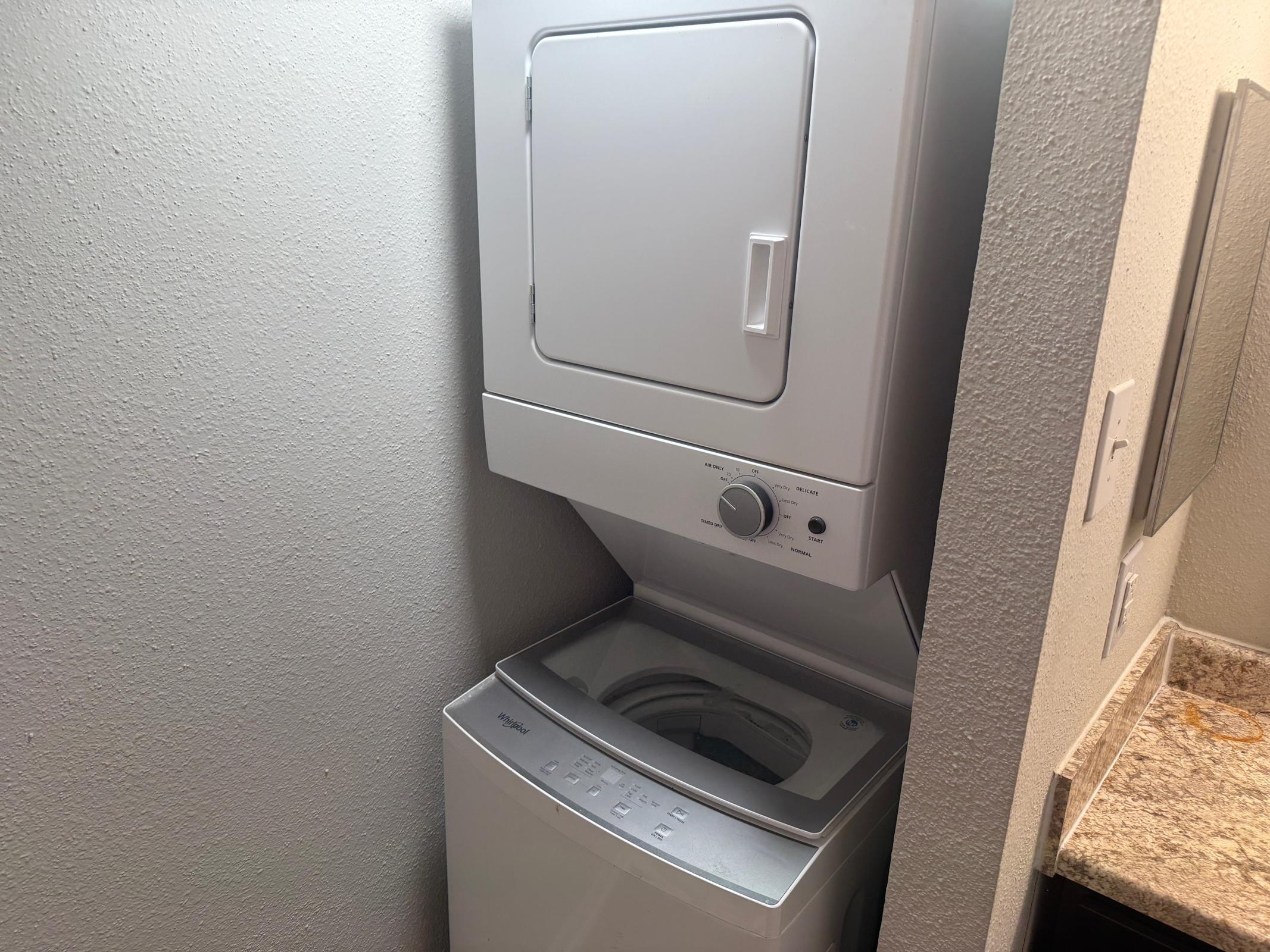 A compact laundry center featuring a stacked washer and dryer unit. The washer is on the bottom with a front-facing opening, while the dryer is above with a control panel on the front. The appliances are situated between a wall and a countertop made of granite, set in a small laundry space.