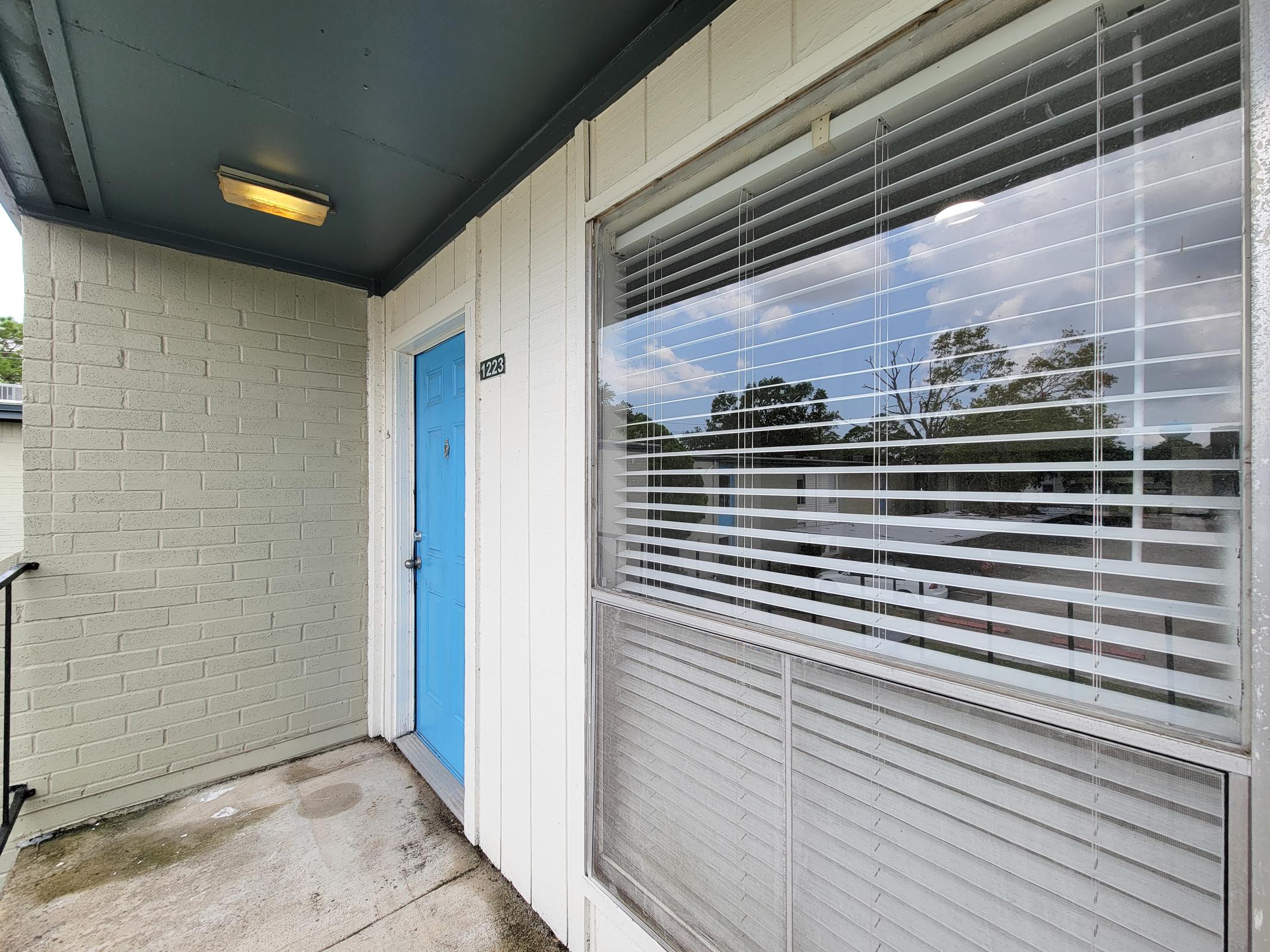 A view of an apartment entrance featuring a blue door, white walls, and a window with blinds. The surrounding area includes trees visible through the window, and the ground is made of concrete, showing a small entrance space. Natural light illuminates the scene.