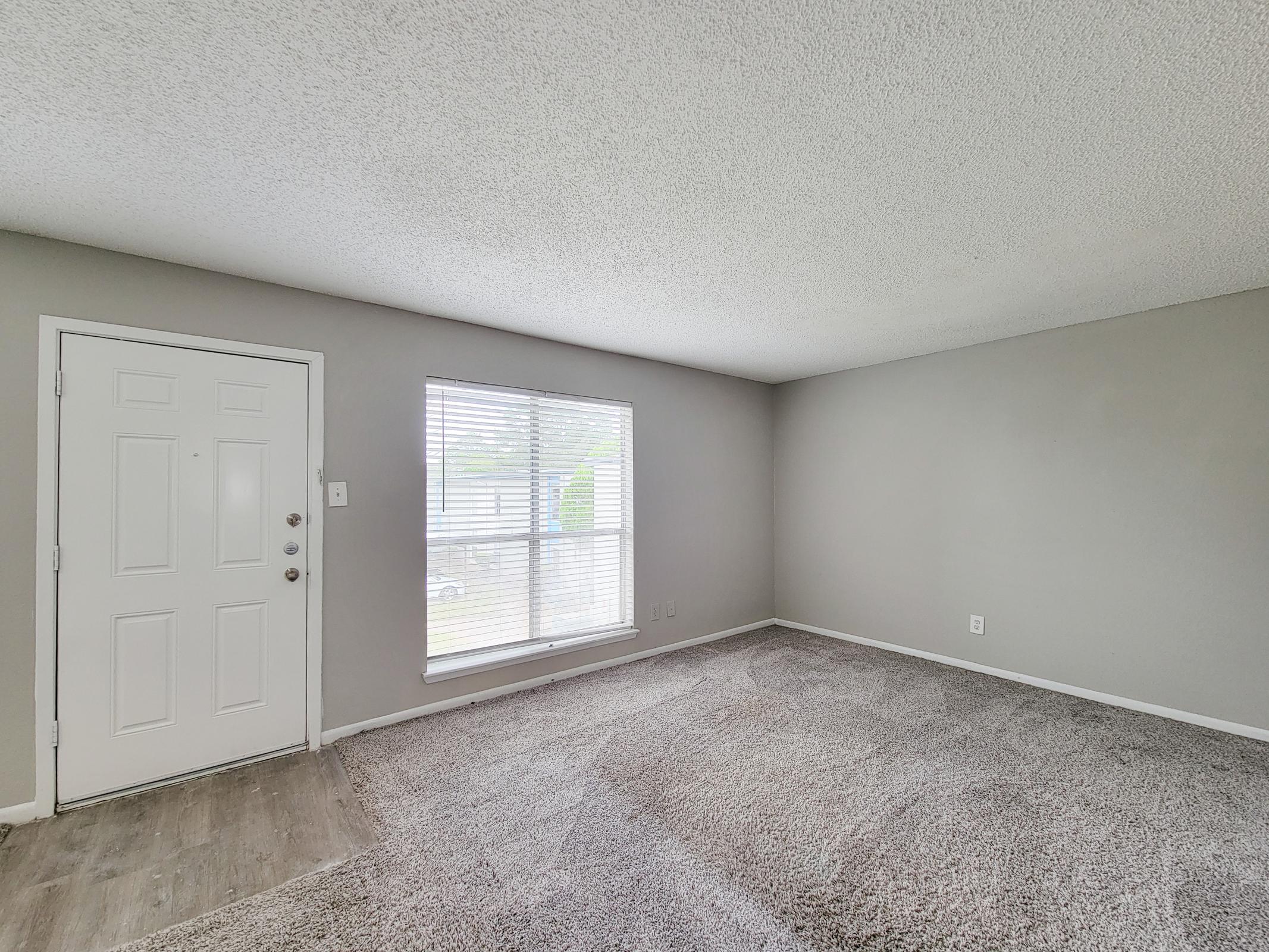 Interior view of a living room with gray walls and carpet. A front door is visible on the left, and a large window with blinds lets in natural light. The room appears spacious and well-lit, with a simple layout and no furniture.