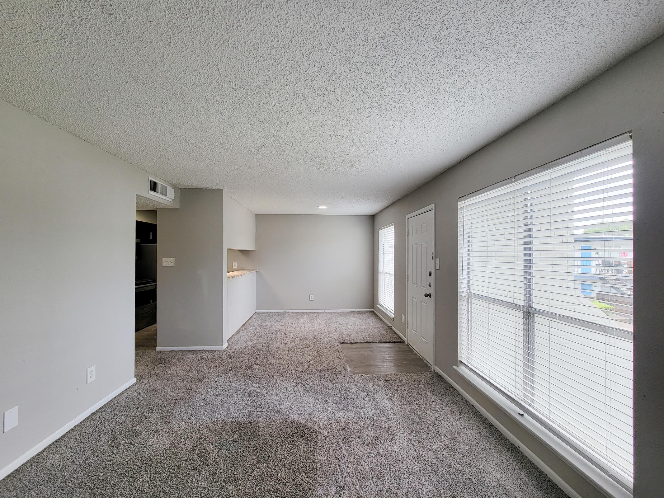 A spacious living area with gray carpet and light gray walls. Large windows allow natural light to fill the room. There's a doorway leading outside and a small kitchenette visible in the background. The area appears clean and inviting, perfect for relaxation or entertaining.