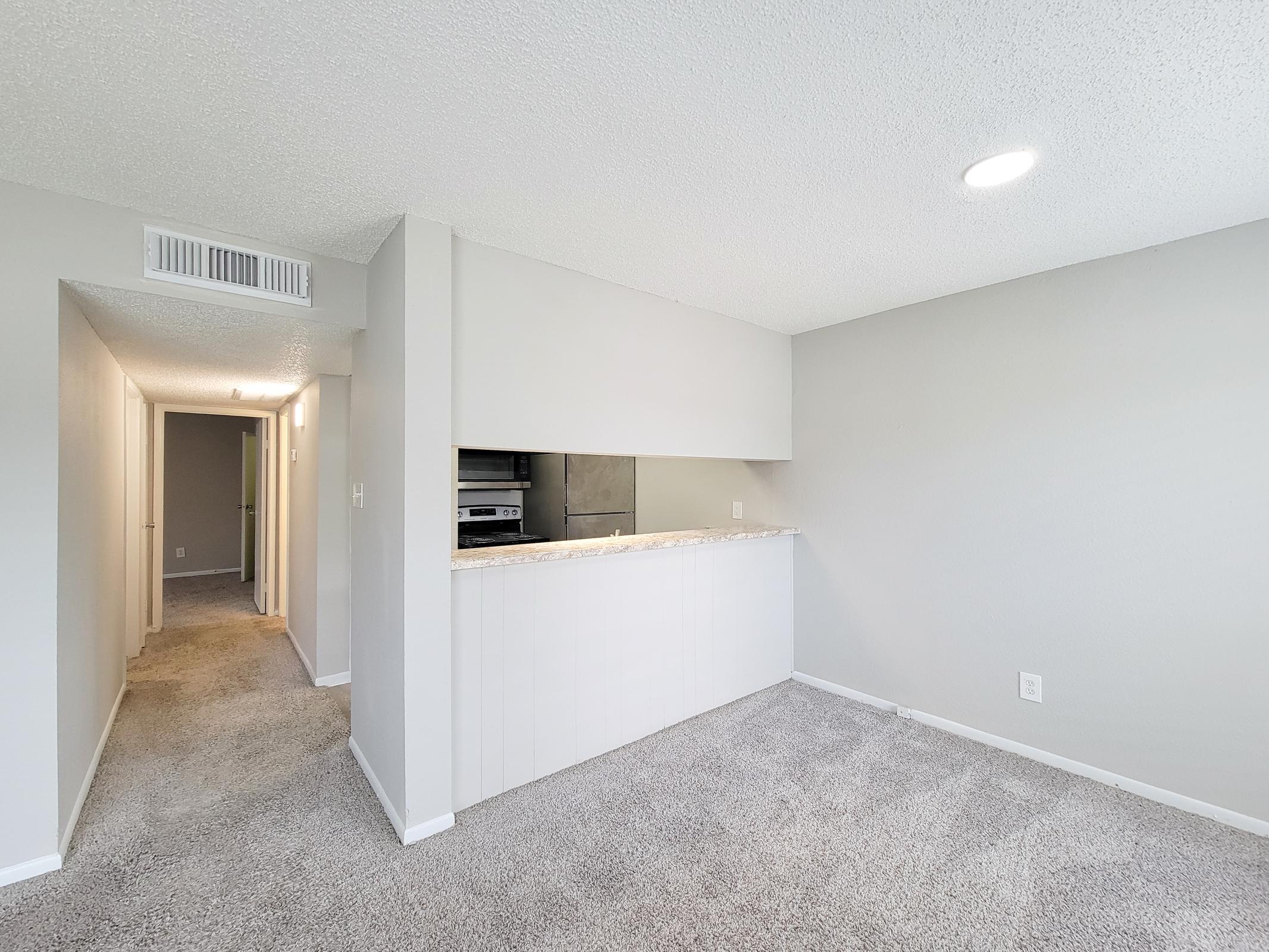 A spacious living area featuring light gray walls and carpet. In the foreground, a partial view of a kitchen bar with a countertop. In the background, a hallway leads to additional rooms, illuminated by natural light from the windows. The overall space is bright and well-maintained.