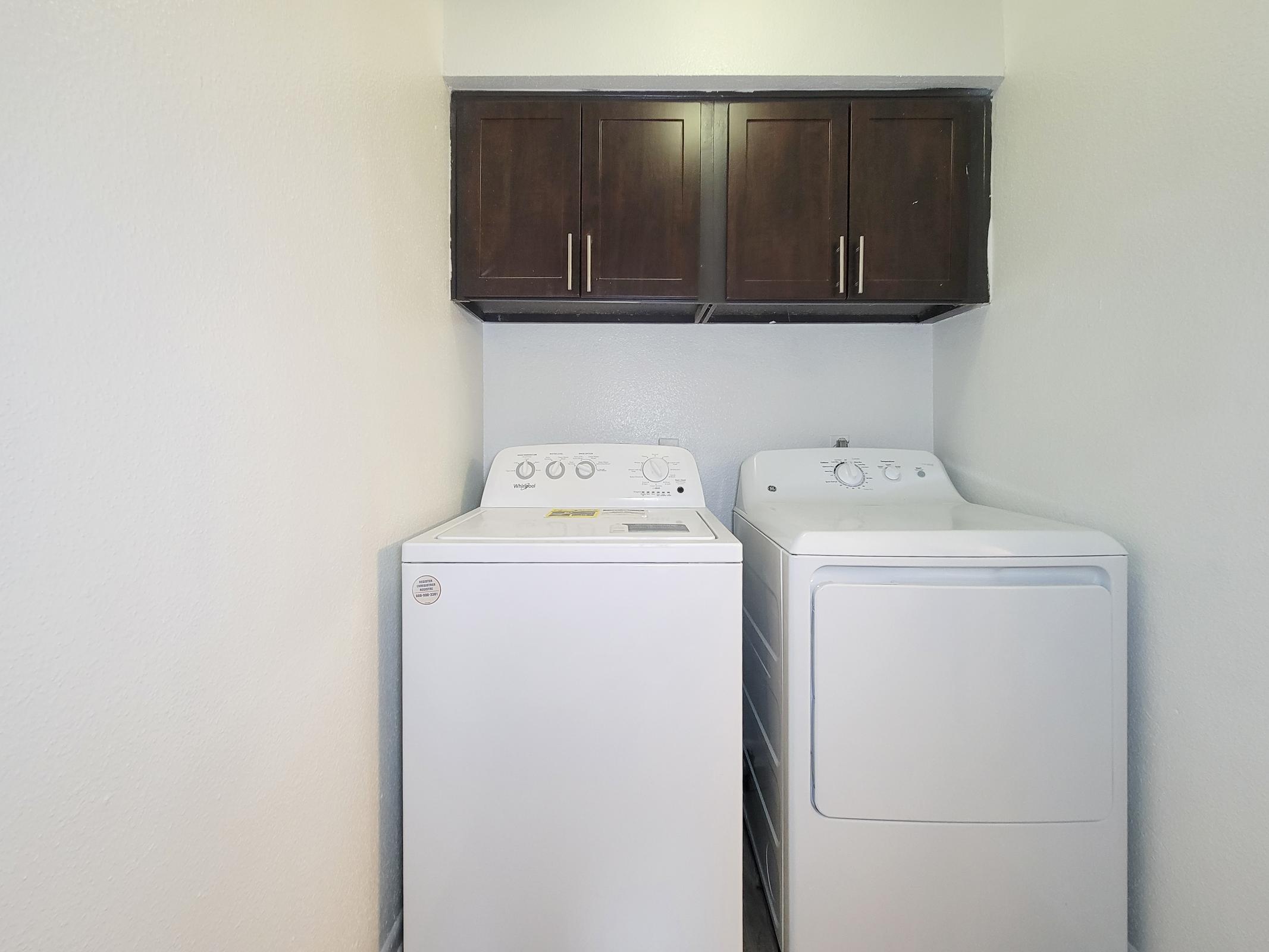 A laundry room featuring a washing machine and a dryer side by side, with dark wooden cabinets above them. The walls are painted white, creating a clean and modern look.