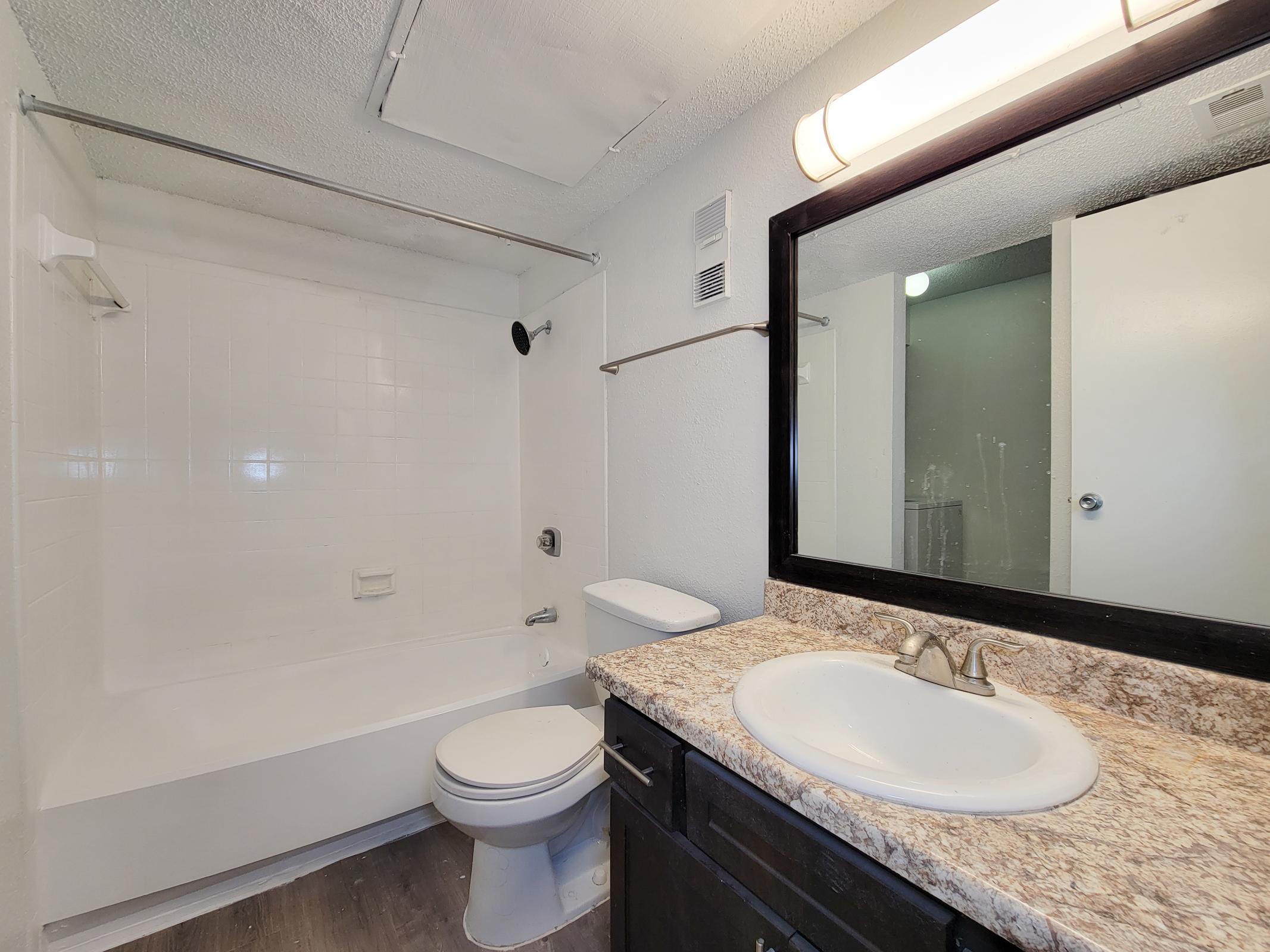 A clean and simple bathroom featuring a white bathtub with a showerhead, a toilet, and a vanity with a sink and mirror. The walls are painted white, and the floor has a dark wood finish. A light fixture is installed above the mirror, providing bright illumination.