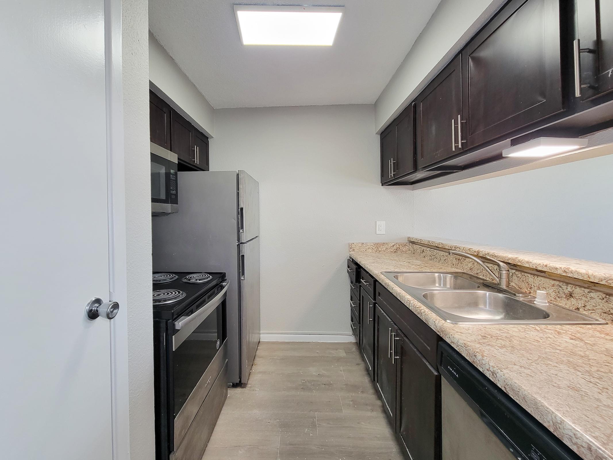 A modern kitchen featuring dark cabinetry, a stainless steel refrigerator, and a stove with an oven. The countertops are made of granite, and there are double sinks. The space is well-lit with recessed ceiling lights and has a neutral color scheme, creating a clean and functional cooking area.