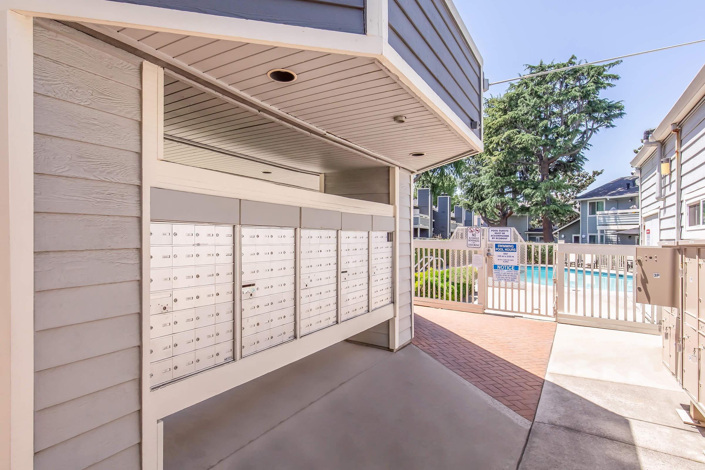 A row of mailboxes situated near a fence and a swimming pool area in an apartment complex. The sky is clear and sunny, with a tree in the background. The architecture features light-colored wood siding and modern design elements.