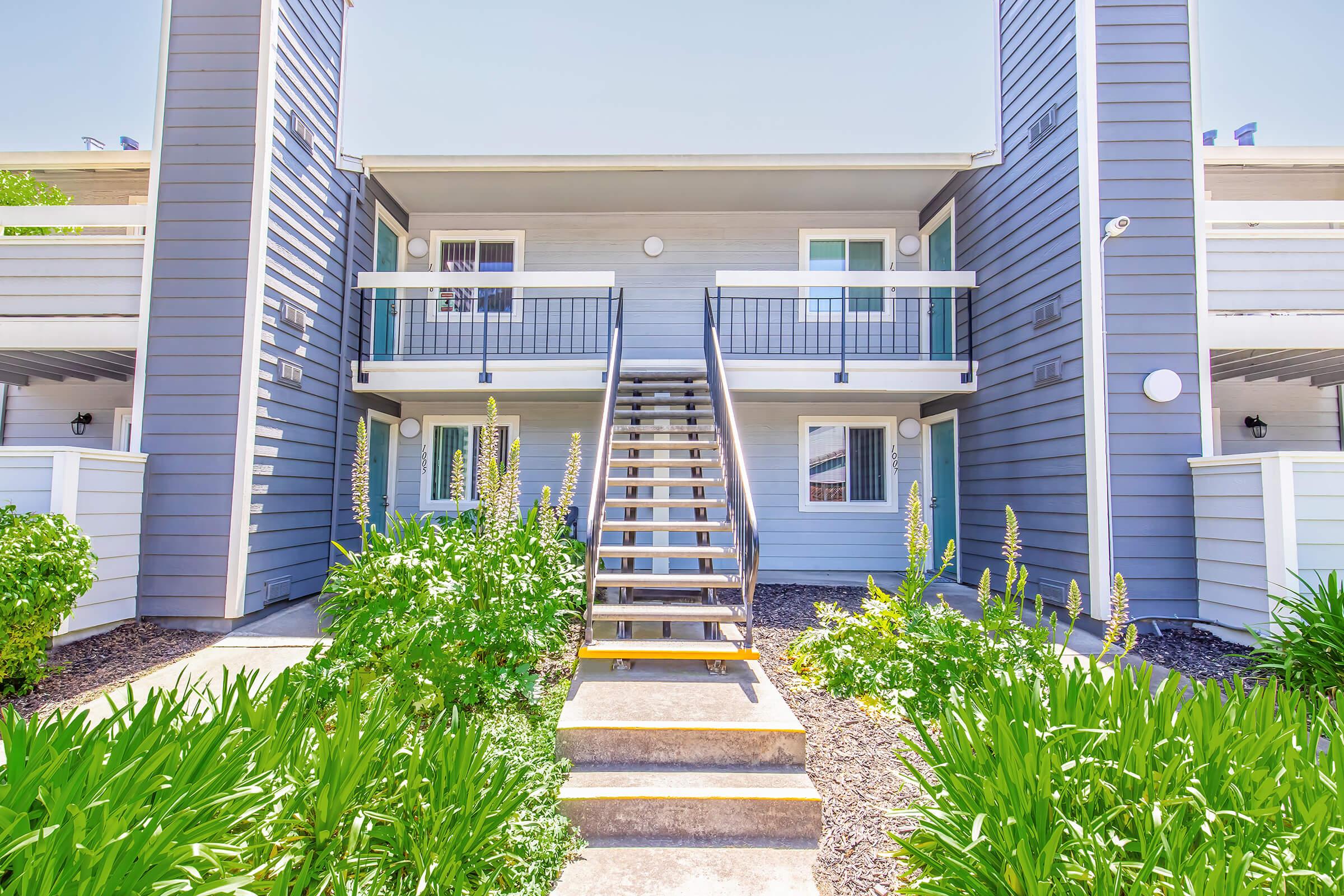 Exterior view of a two-story apartment building featuring a staircase leading to the upper level. The building has gray siding, balconies, and windows with railings. Lush green plants and landscaping adorn the entrance, creating an inviting atmosphere. Clear blue sky in the background adds to the bright ambiance.