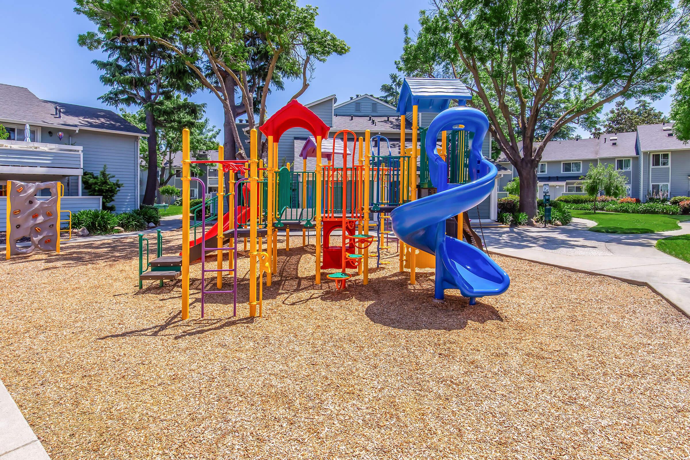 Colorful playground equipment with slides, climbing structures, and swings, surrounded by a sandy play area and trees. Brightly colored structures in red, blue, and yellow invite children to play in a sunny outdoor setting. Nearby houses and greenery provide a welcoming, family-friendly atmosphere.