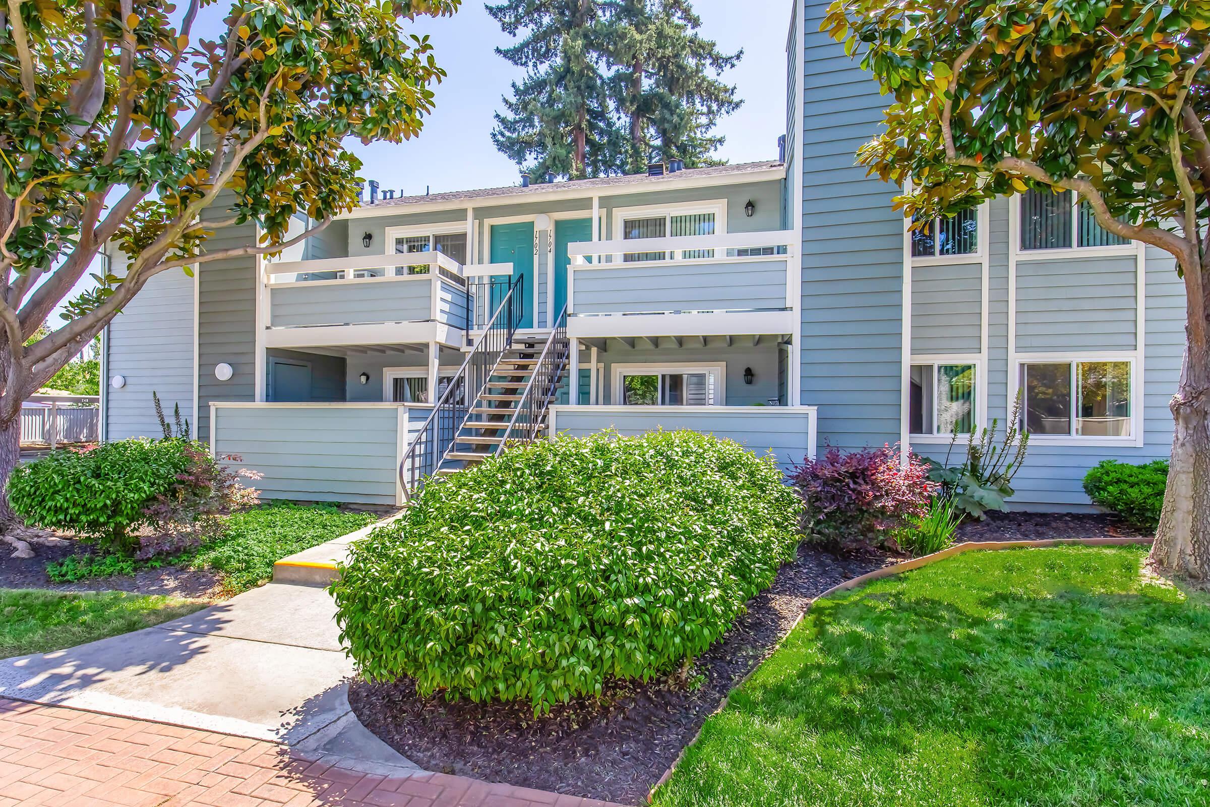 Two-story apartment building with blue siding, featuring multiple balconies and white railings. The entrance has a set of stairs leading up to the doors. Surrounding the building are well-maintained green shrubs and grass, along with trees adding shade to the area. A brick walkway leads to the building's entrance.