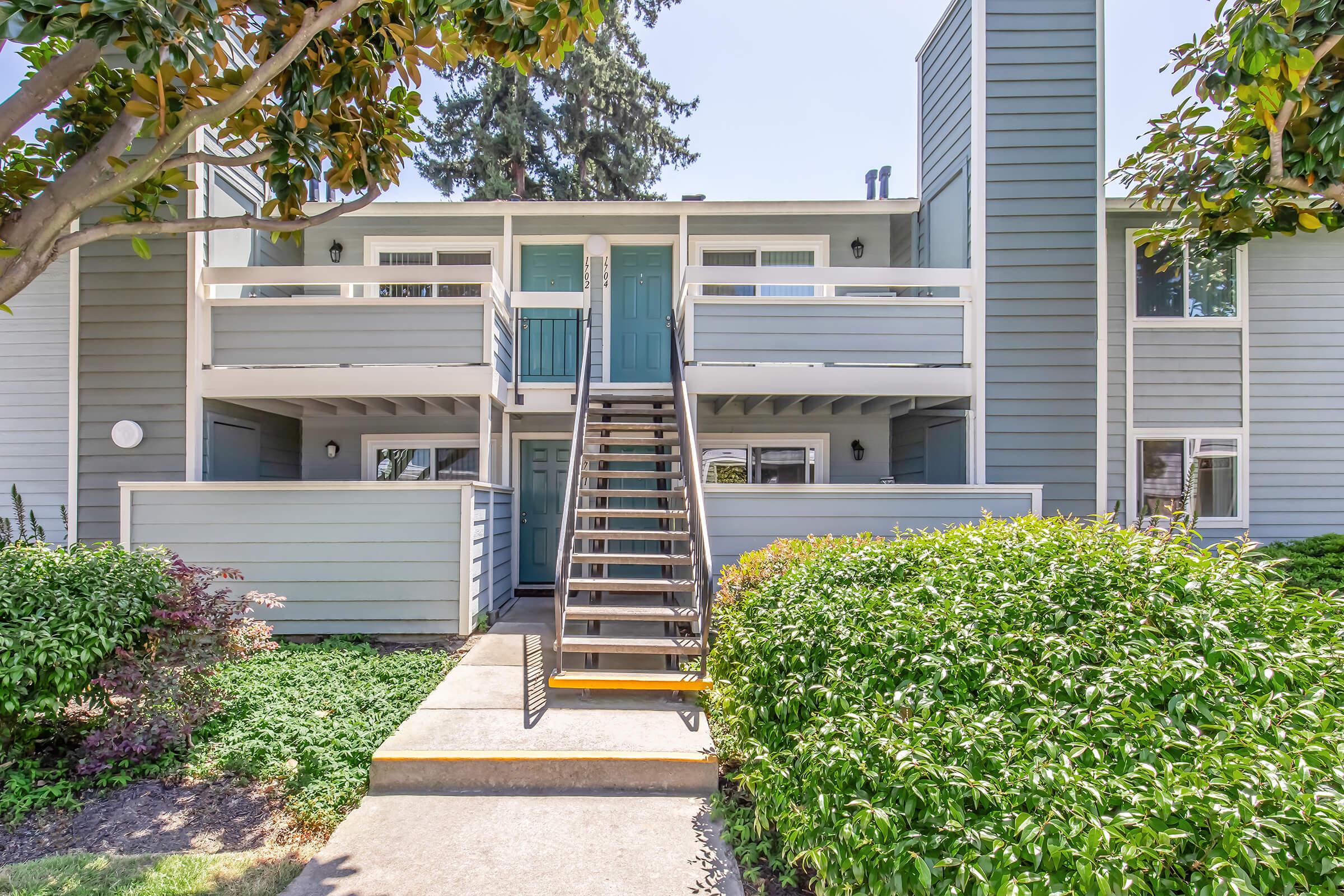 A light blue apartment building features a two-story design with a central staircase leading to the second-floor entrances. Each level has a balcony with white railings. Lush greenery surrounds the walkway, and the scene is bright under clear blue skies.