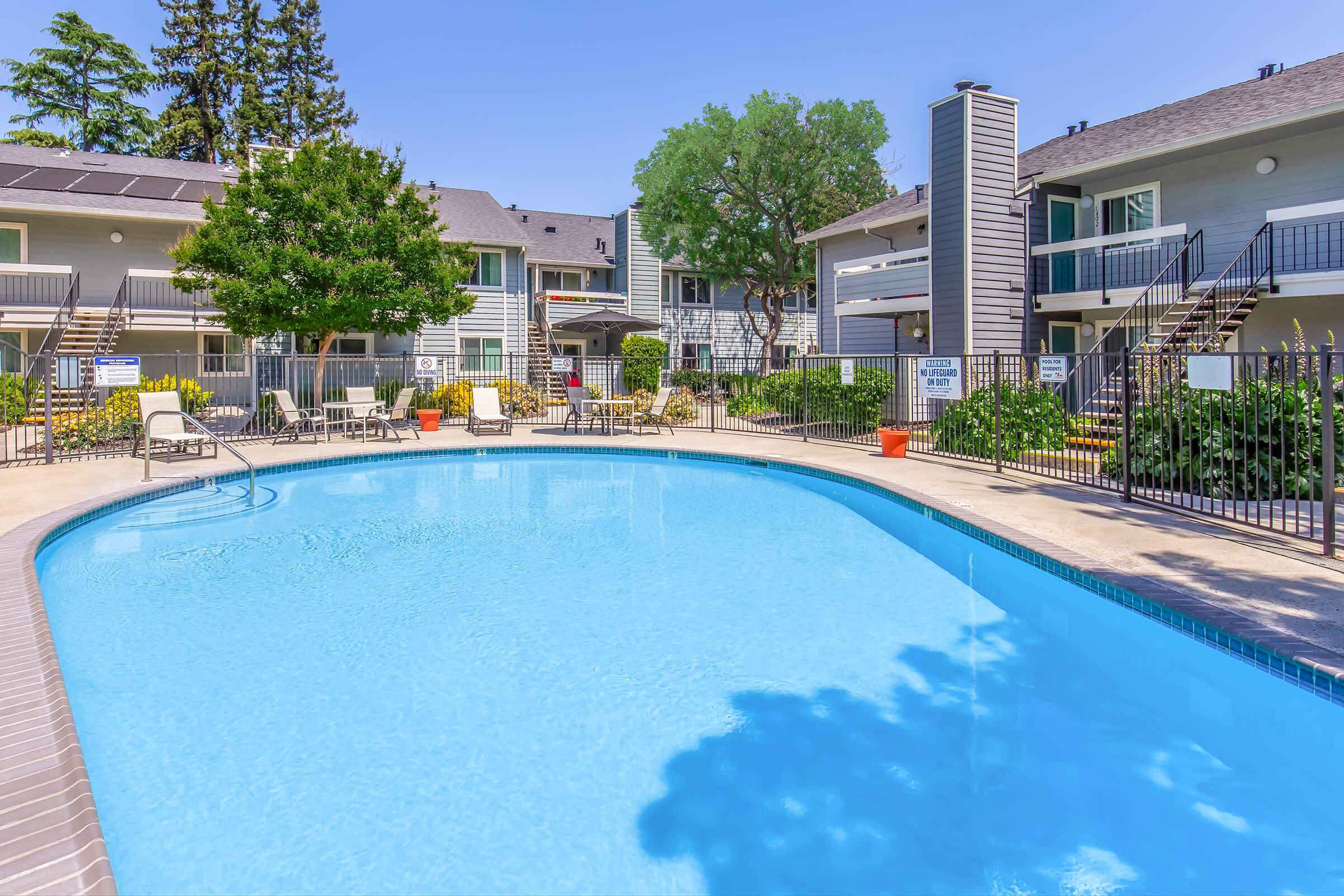 A clean, blue swimming pool surrounded by lounge chairs and greenery, located in a residential complex. Several apartment buildings are visible in the background, under a clear blue sky. The area is well-maintained and inviting, ideal for relaxation or leisure activities.