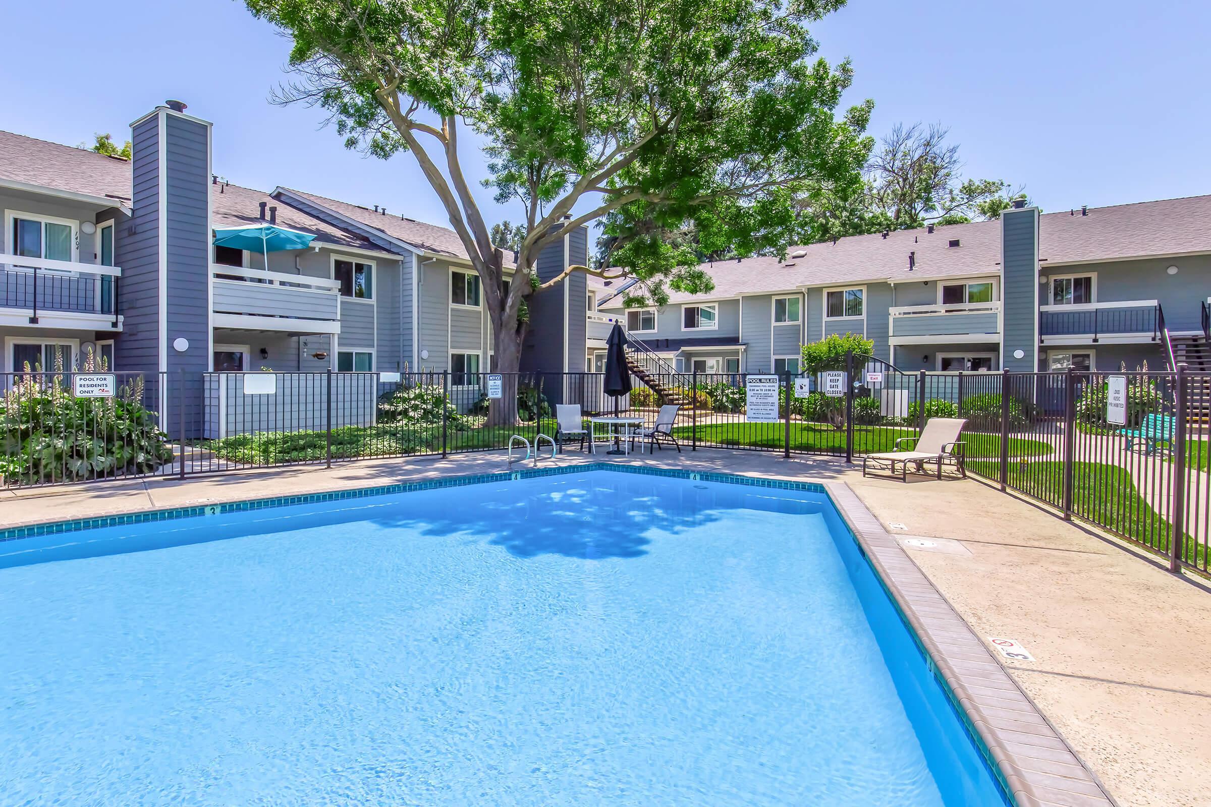 A clear blue swimming pool surrounded by a fenced area, with lounge chairs placed nearby. In the background, there are two-story apartment buildings with balconies, greenery, and trees. The scene is bright and inviting, suggesting a relaxing outdoor space.