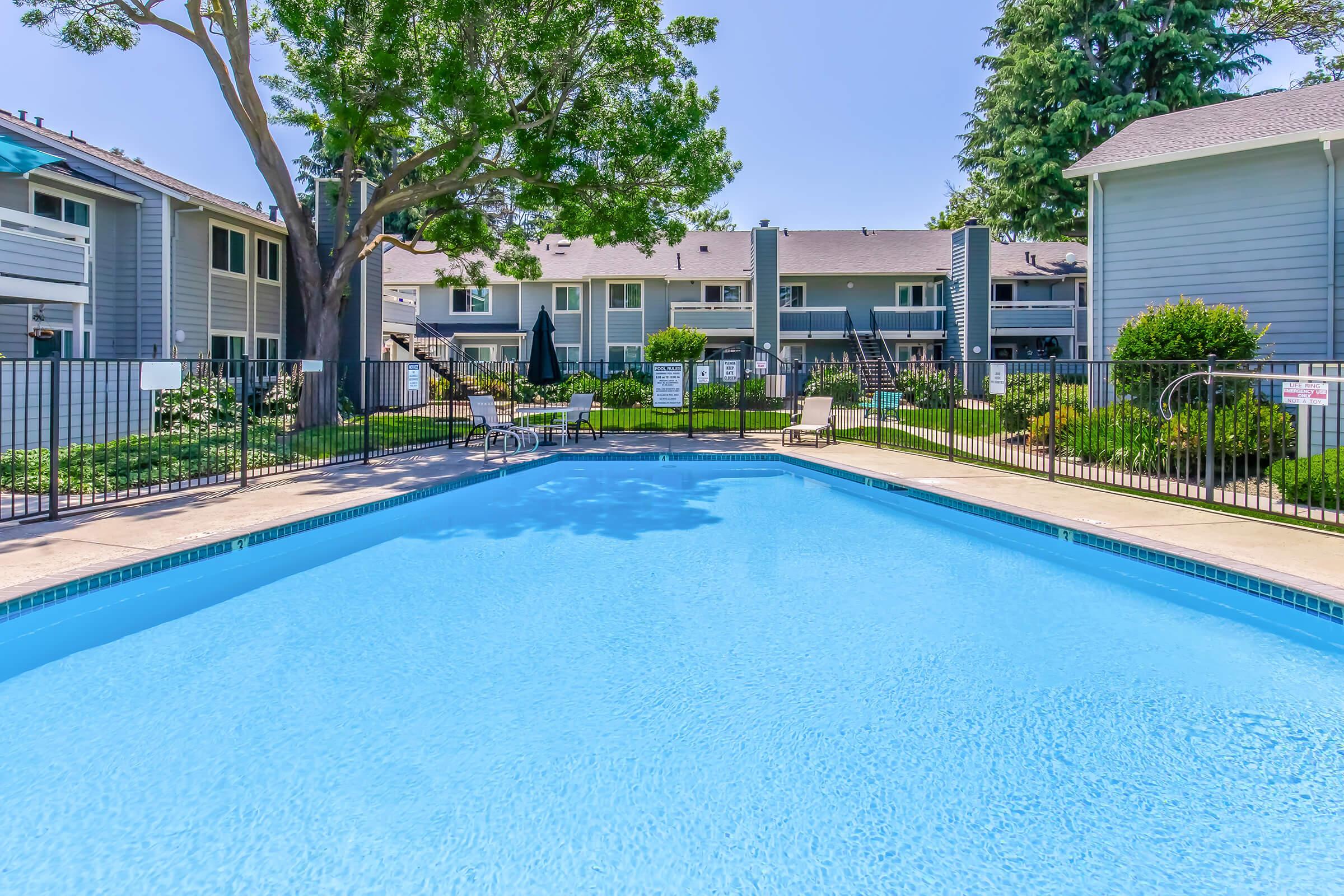 A clear blue swimming pool with gentle ripples reflects the surrounding area. Lush greenery and lounge chairs are positioned around the pool, while two-story apartments are visible in the background under a bright blue sky. The scene conveys a serene and inviting atmosphere.
