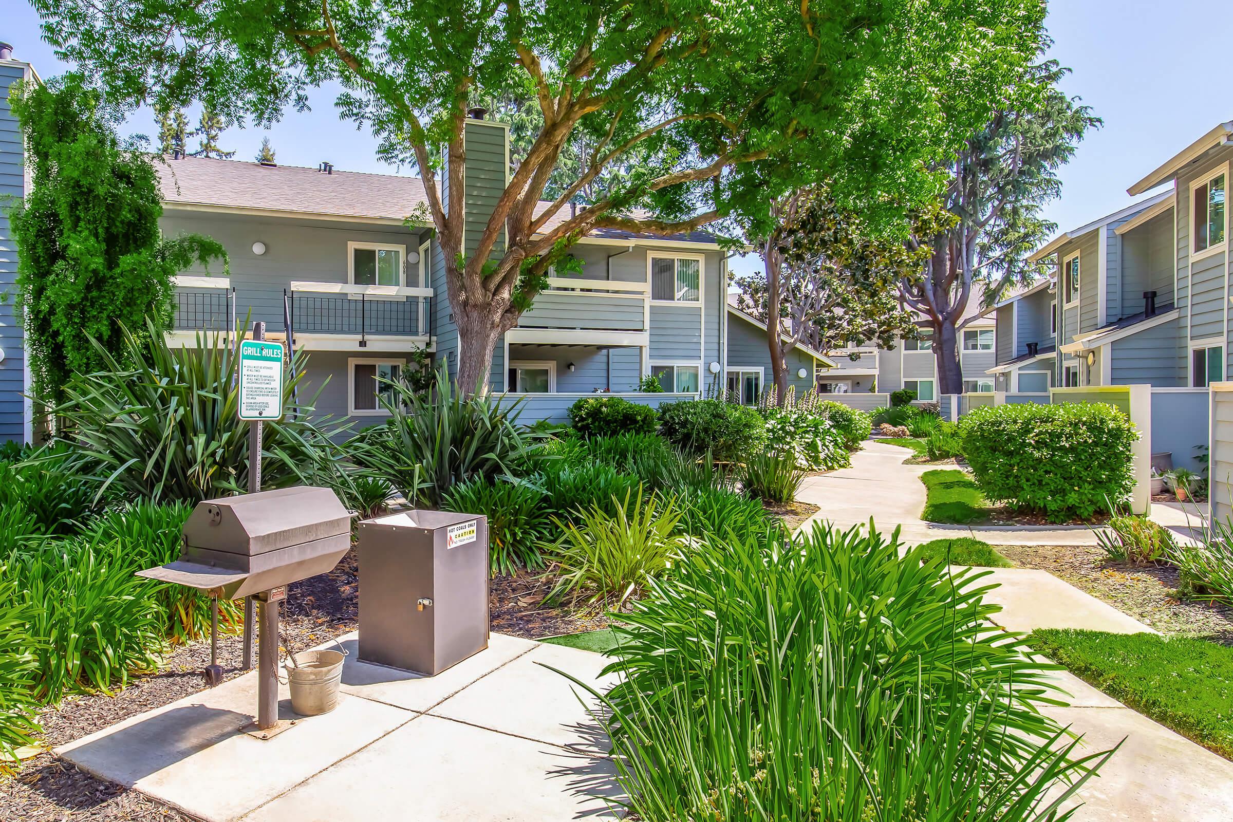 A landscaped pathway leads through a green courtyard featuring well-maintained shrubs and plants. There are two residential buildings in the background, and a small barbecue grill stands to the left, creating a welcoming outdoor environment.