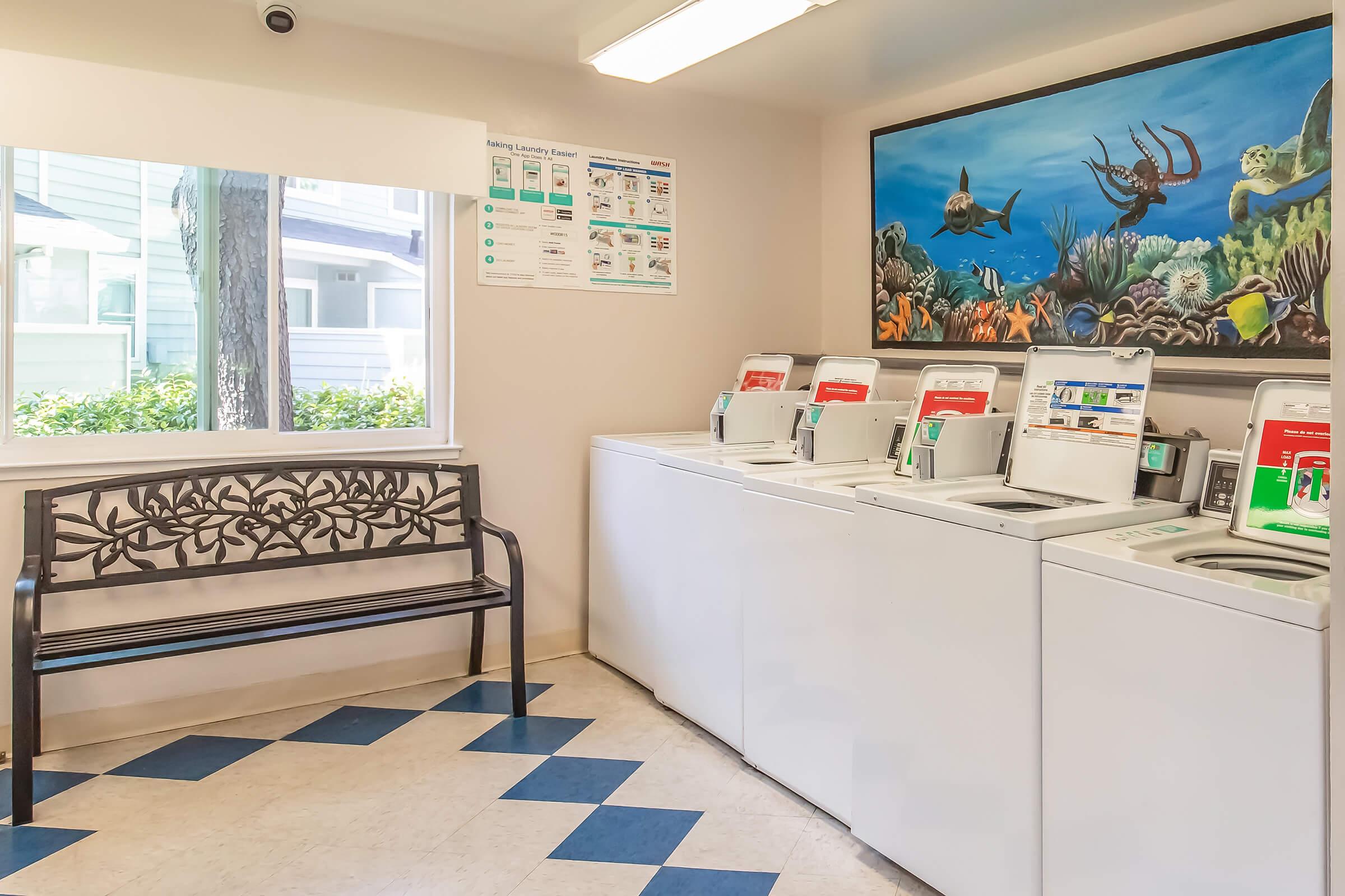 Interior view of a laundry facility featuring a seating area with a decorative bench. There are multiple washing machines with digital displays. A colorful underwater-themed mural adorns the wall, depicting marine life such as fish and corals. Natural light comes in through a window.