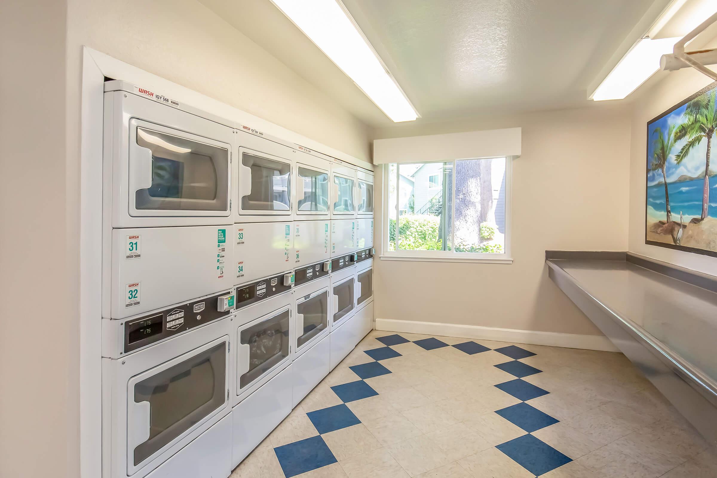 A clean, well-lit laundry room featuring stacked washer and dryer units along one wall. A large window provides natural light, and there's a beach-themed mural on the adjacent wall. The floor is tiled with a diamond pattern in blue and beige.