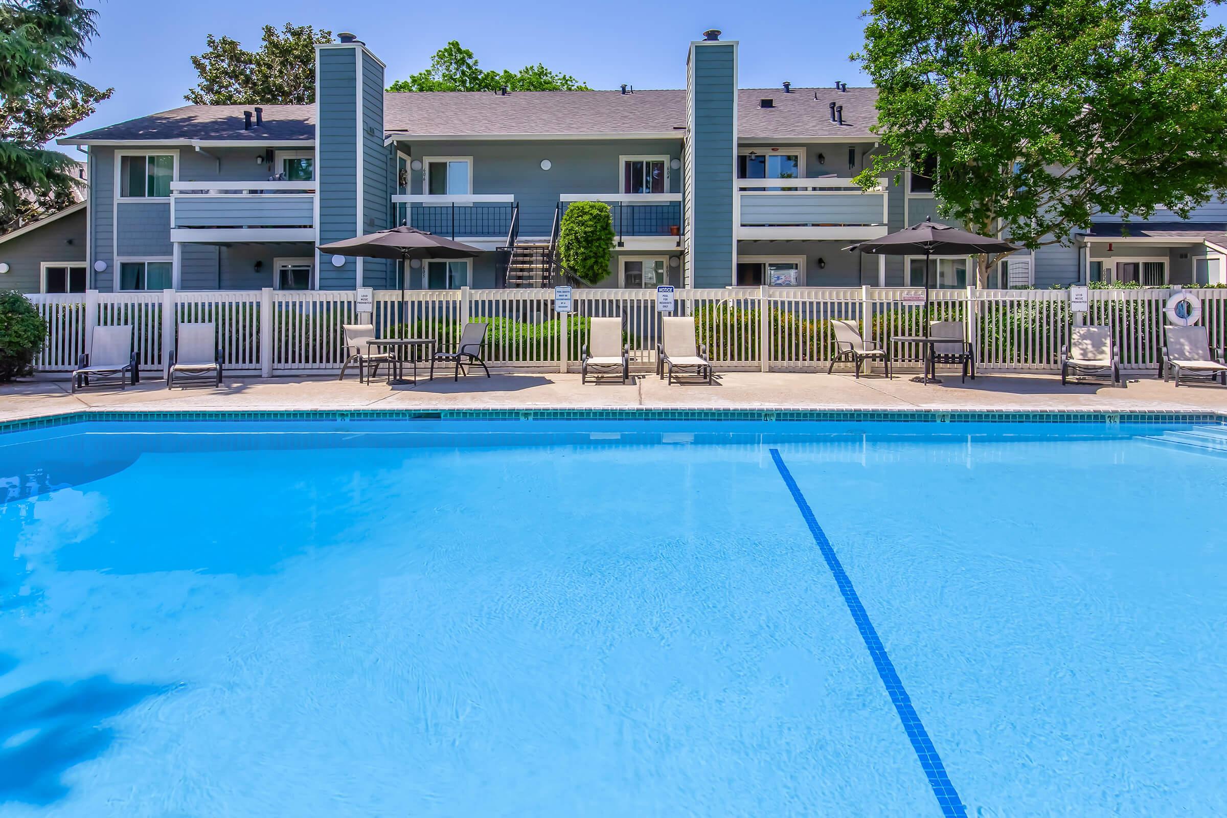 A clear blue swimming pool surrounded by lounge chairs and umbrellas, with apartment buildings in the background. Lush green trees and grass enhance the outdoor area, creating a relaxing recreational environment.
