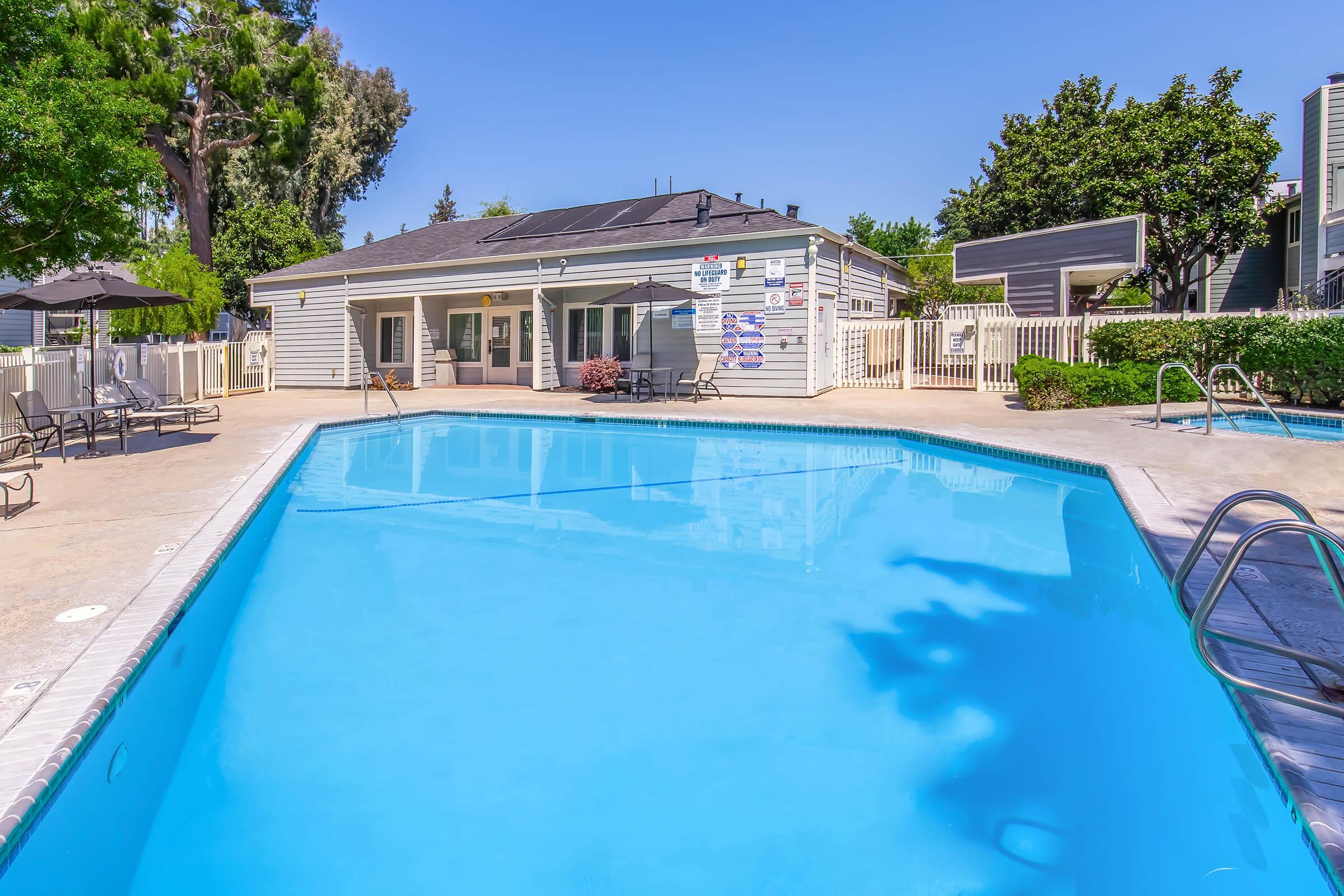 A bright blue swimming pool with clear water, surrounded by lounge chairs and umbrellas. In the background, there is a building with outdoor amenities, set against a sunny sky and greenery. The scene conveys a relaxing and inviting atmosphere.