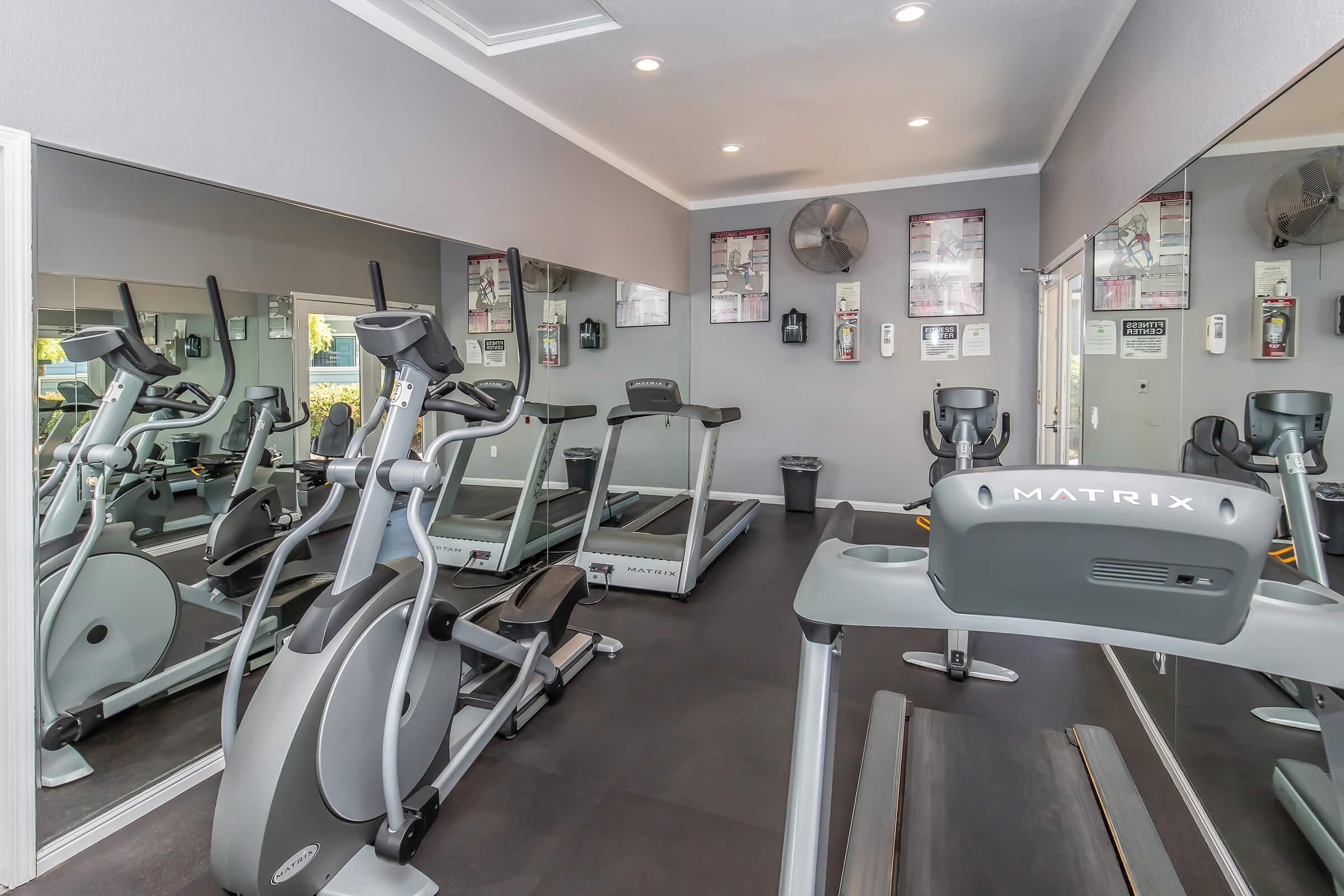 A modern gym interior featuring multiple exercise machines, including a treadmill and an elliptical, set against a backdrop of mirrors and wall-mounted fitness posters. Bright lighting and a clean, spacious design create an inviting atmosphere for workouts.