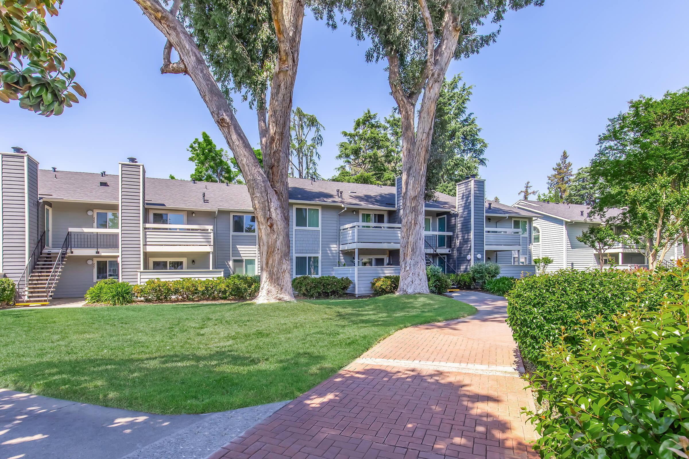 A residential complex featuring two-story buildings surrounded by lush greenery and large trees. A paved pathway winds through a well-maintained lawn, leading to the entrances of the apartments. The sky is clear and blue, creating a bright and inviting atmosphere.