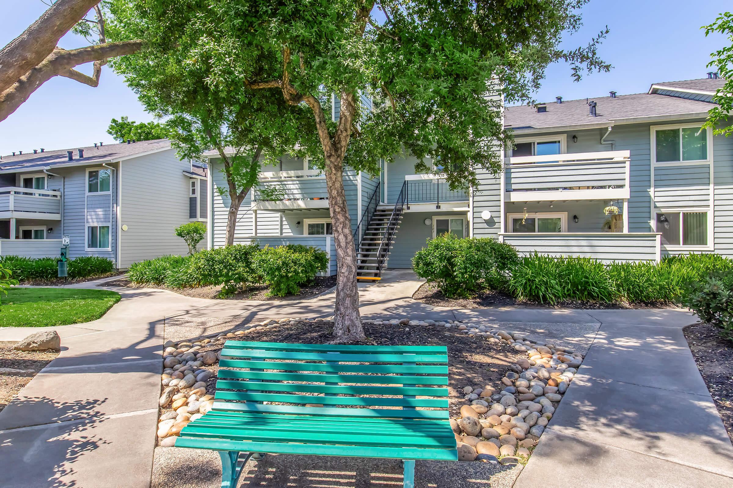 A vibrant green bench sits in a landscaped courtyard surrounded by trees and shrubbery, in front of a multi-unit residential building. The sunny day enhances the inviting atmosphere, showcasing paths and decorative rocks that add to the tranquil setting.